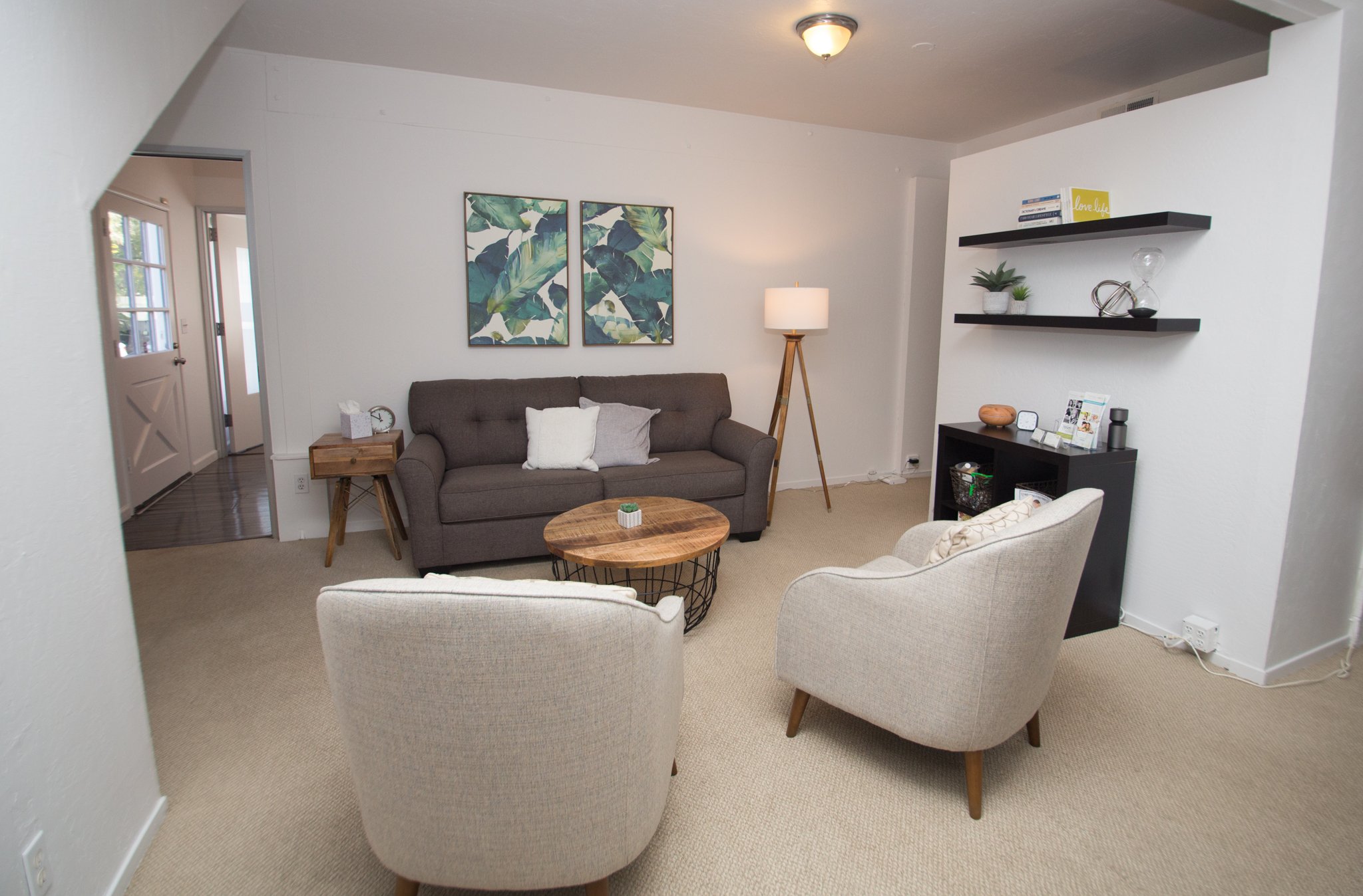 Living room with beige carpet, two upholstered chairs, a gray sofa with pillows, a wooden coffee table, a tall floor lamp, wall art with green leaves, and wall shelves with books and decorative items.