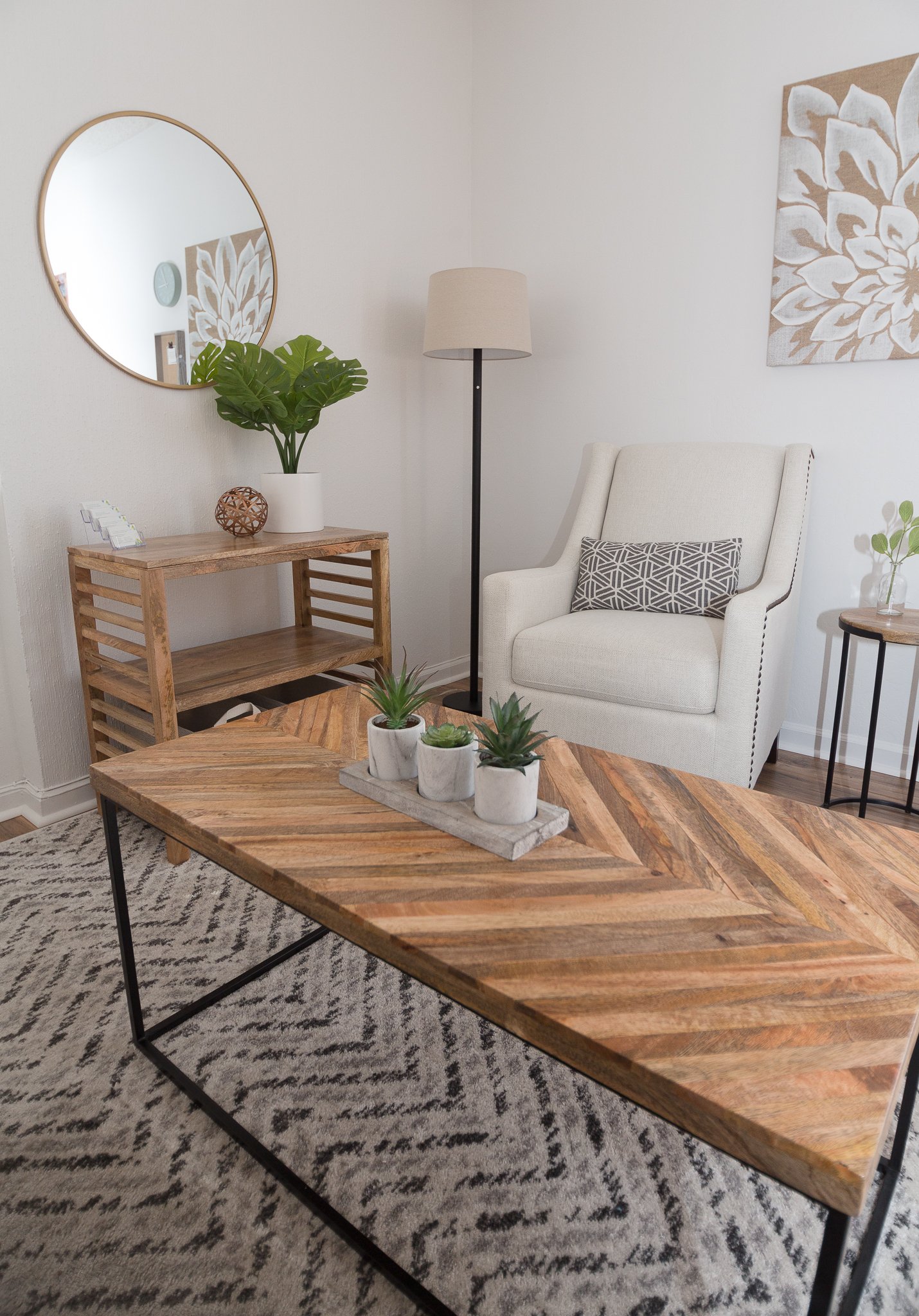 Living room with a wooden table, a white armchair with a geometric pillow, a tall floor lamp, a wooden console with a plant and decorative object, a round mirror on the wall, and a framed floral art piece.