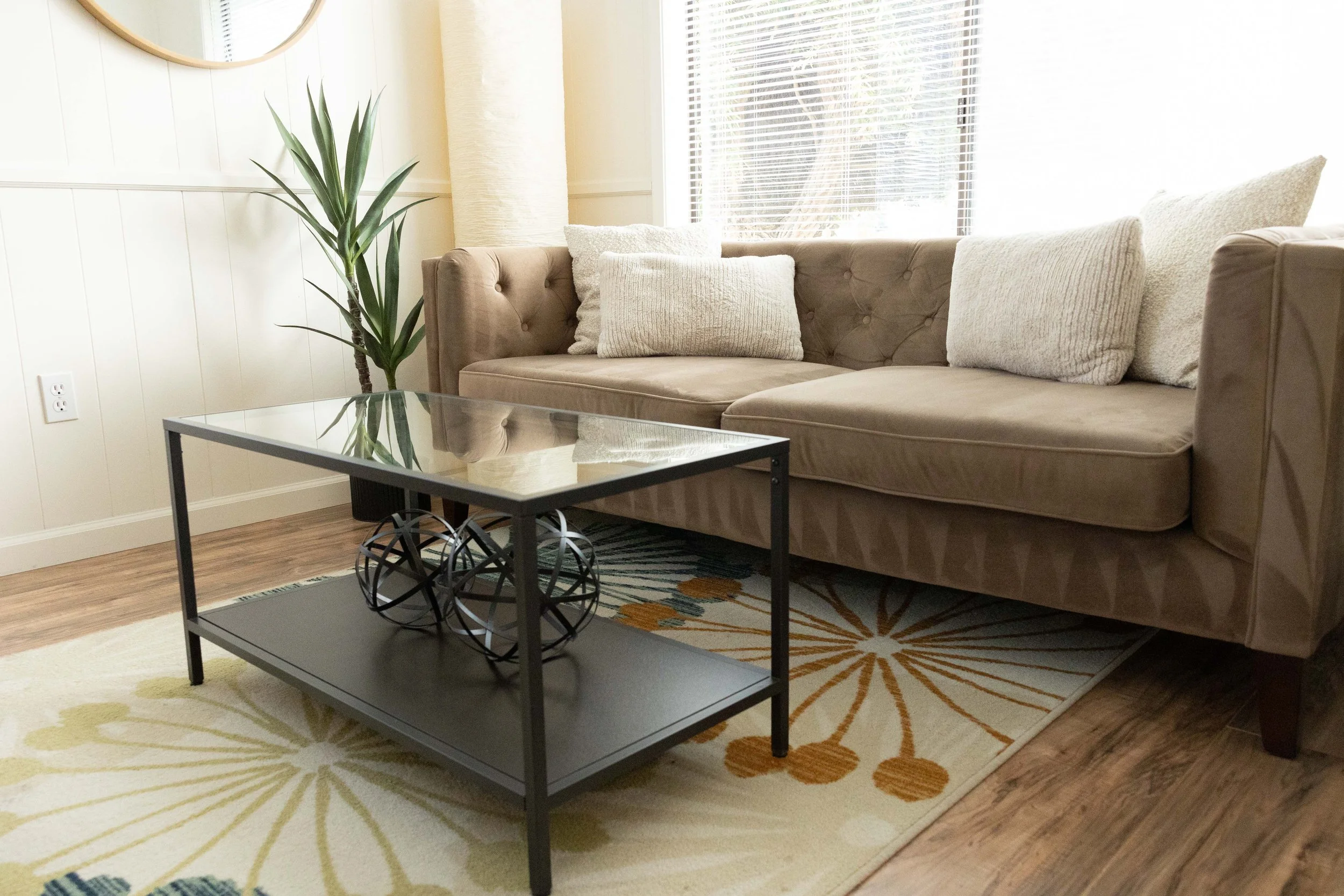 Living room with beige sofa, white cushions, large window with blinds, potted plant, glass-top coffee table, patterned rug, and wall mirror.