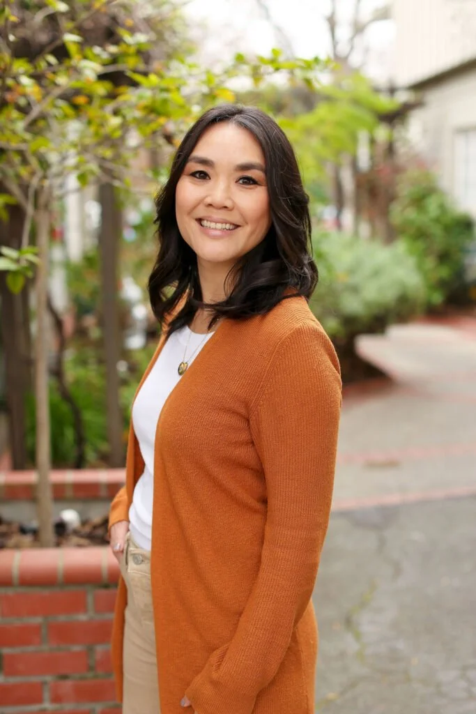 A woman standing outdoors on a sidewalk with trees and bushes in the background, smiling and looking at the camera, wearing a white shirt, beige pants, and an orange cardigan.