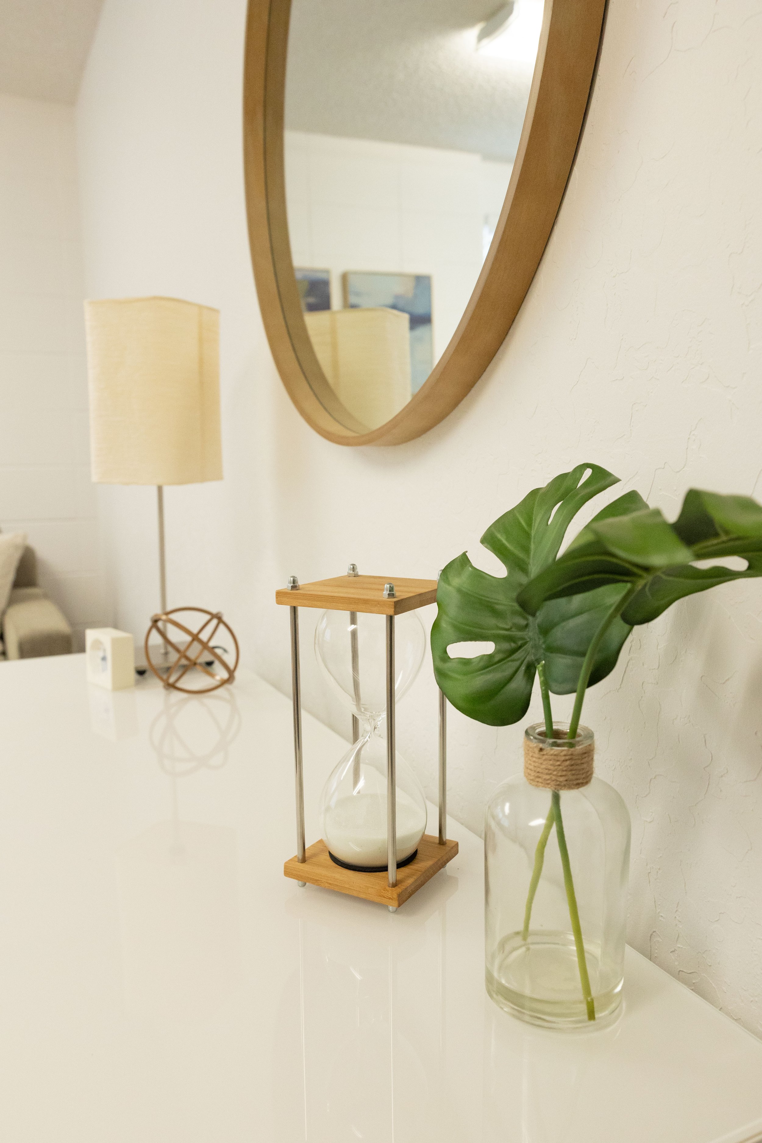 A white table with a small wooden and glass decorative piece, a lamp with a beige shade, a round mirror on the wall, and a potted plant with large green leaves in a glass vase.