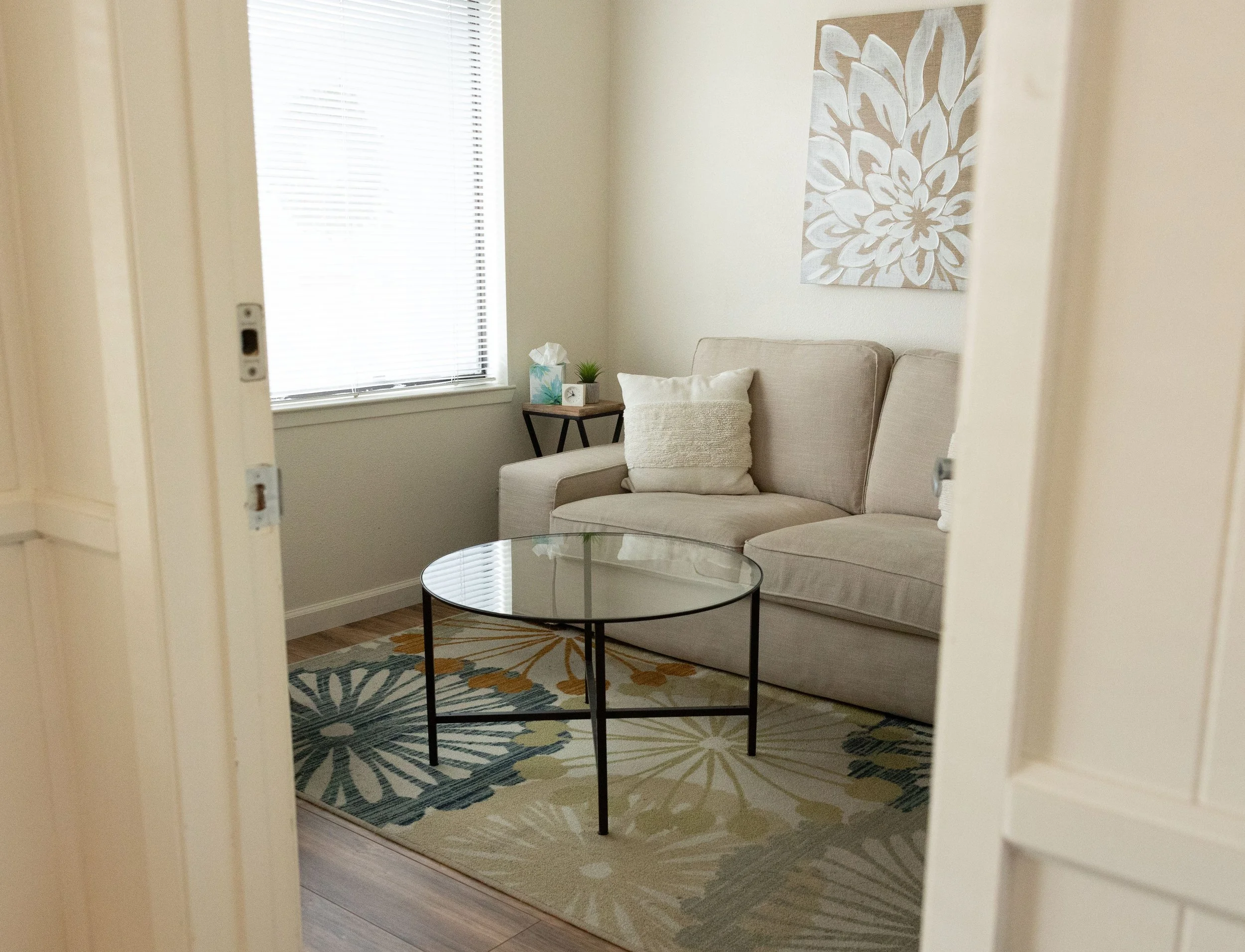 A cozy living room corner with a beige sofa, a small wooden side table with plants and tissue box, a round glass coffee table, a colorful patterned rug, and a large floral painting on the wall near a window with blinds.