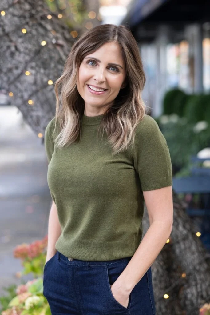 A smiling woman with wavy brown hair in an outdoor setting, wearing a green short-sleeve sweater and dark blue jeans.
