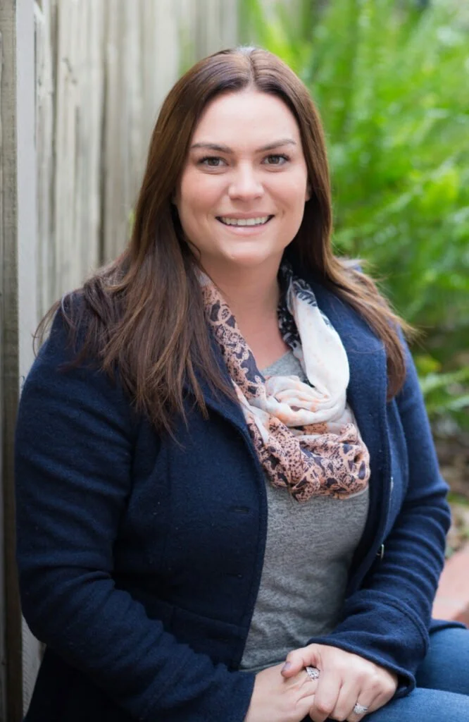 Portrait of a smiling woman with brown hair, wearing a navy blazer, gray shirt, and patterned scarf, standing outdoors near a wooden fence and green plants.