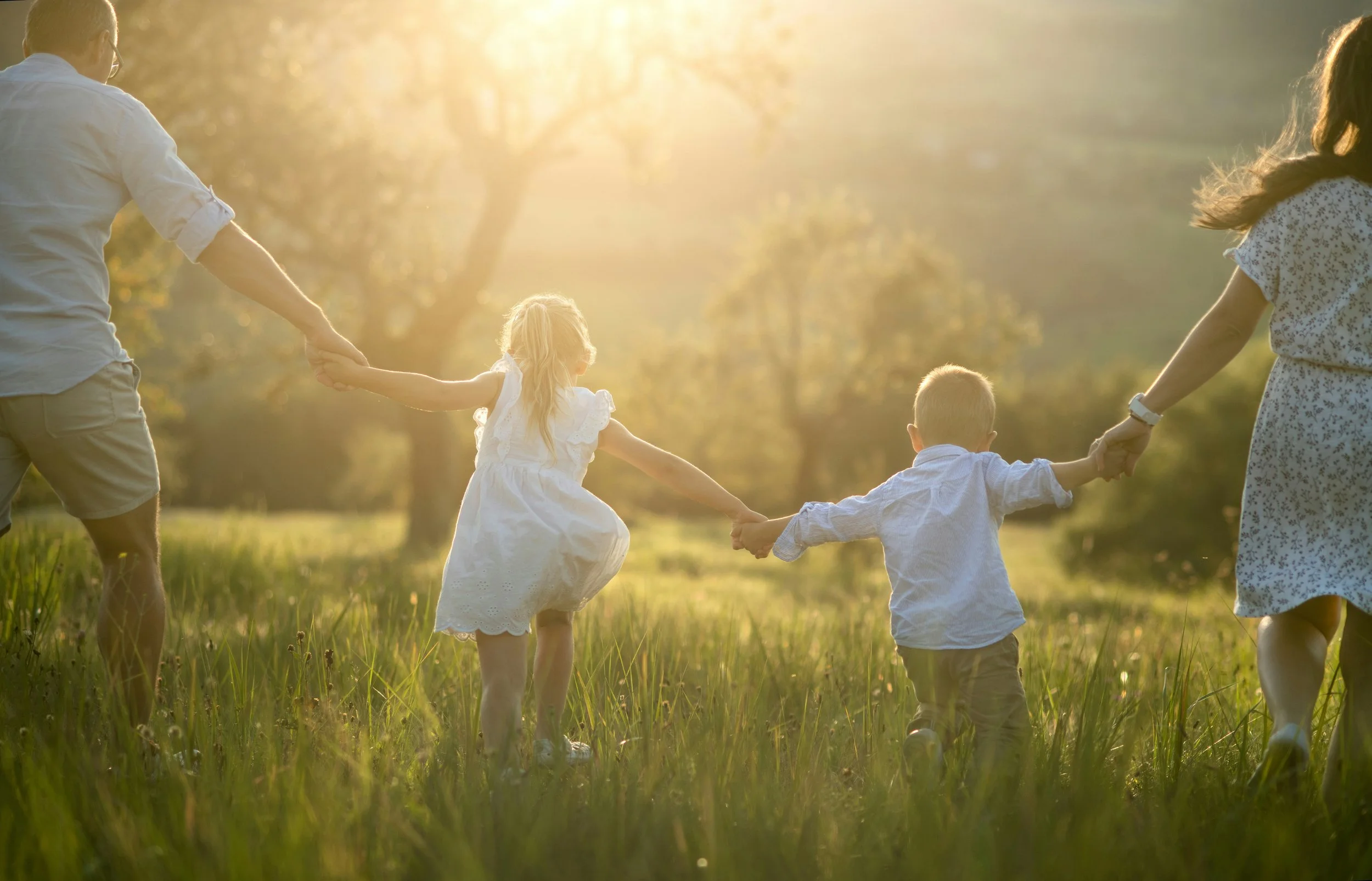A family of four holding hands and playing outdoors in a grassy field during sunset.