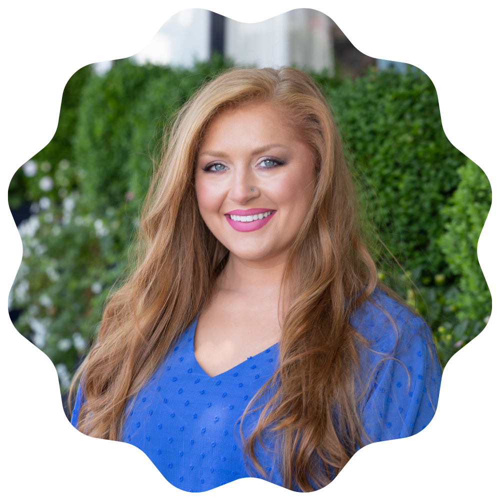 A woman with long wavy red hair, wearing a blue top, smiling outdoors with greenery in the background.