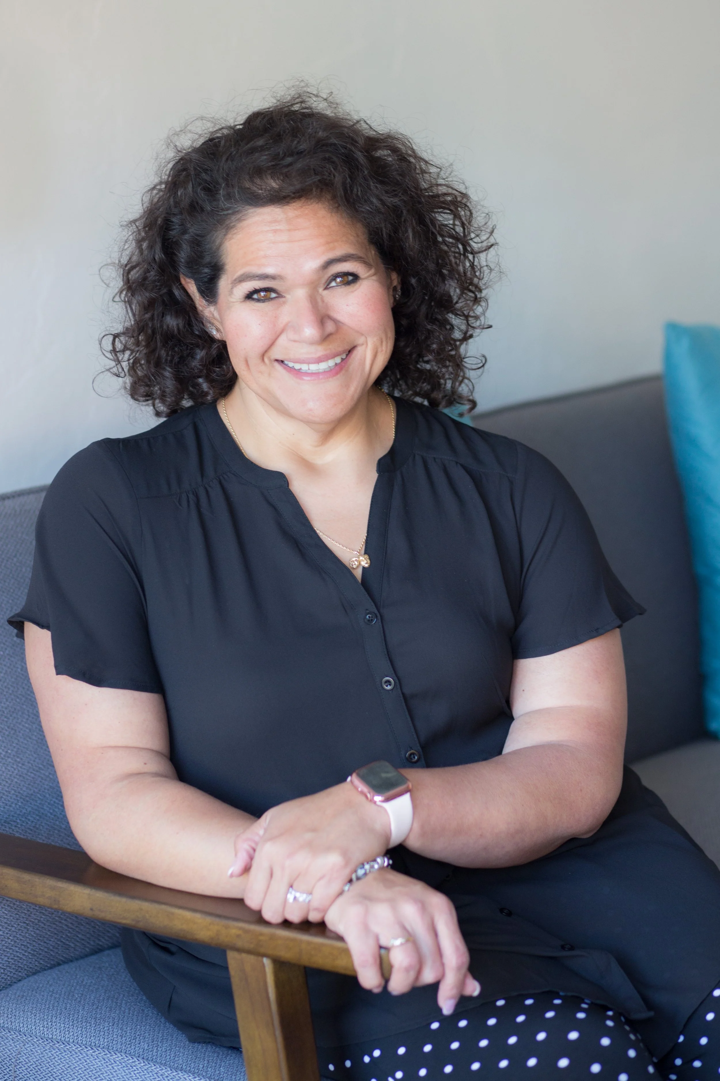 A woman with curly dark hair smiling, wearing a black top, sitting on a gray chair with a wooden armrest, holding a smartwatch, and wearing jewelry including rings, earrings, a necklace, and a bracelet.