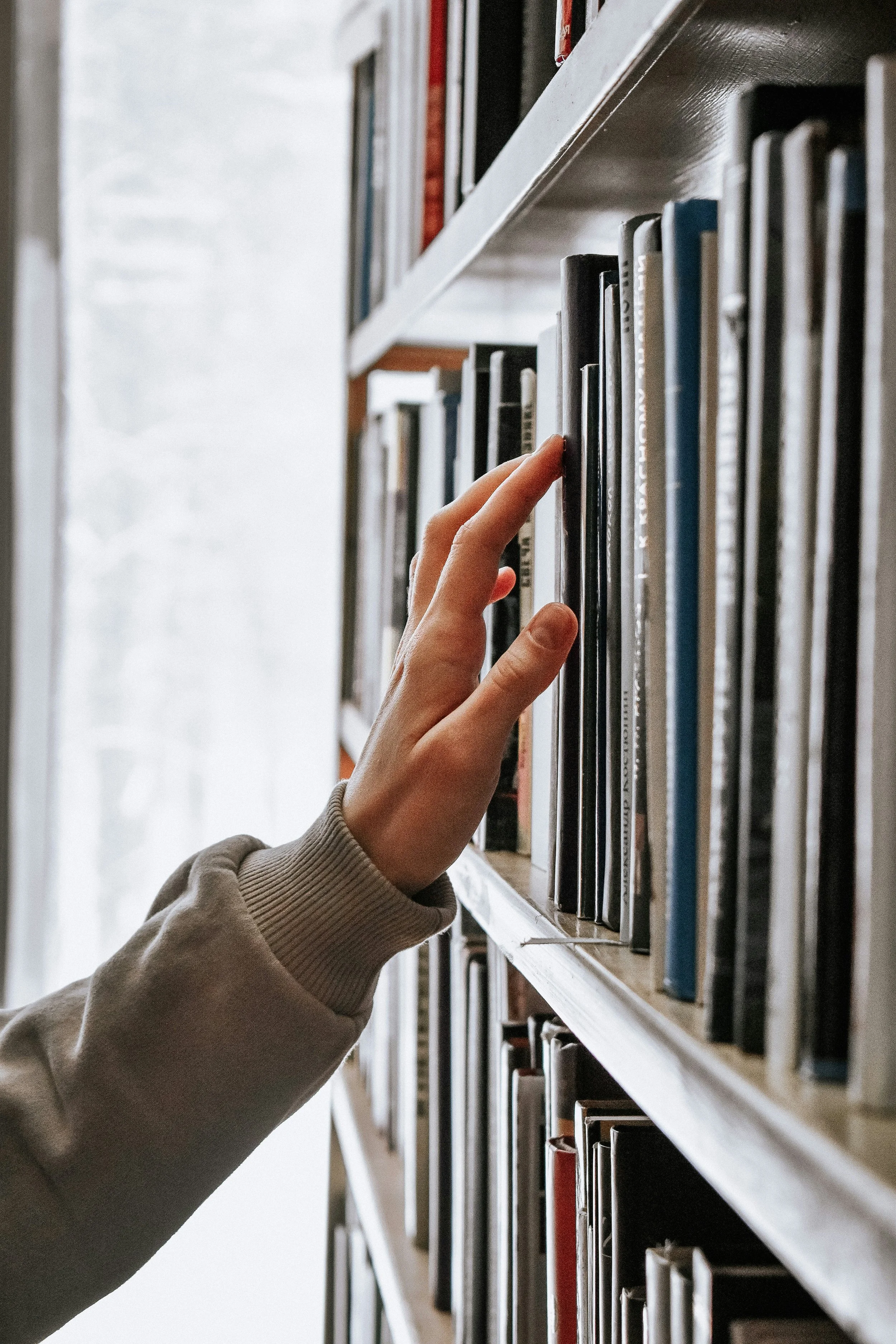 A person's hand reaches for a book on a bookshelf filled with books in a library or bookstore.