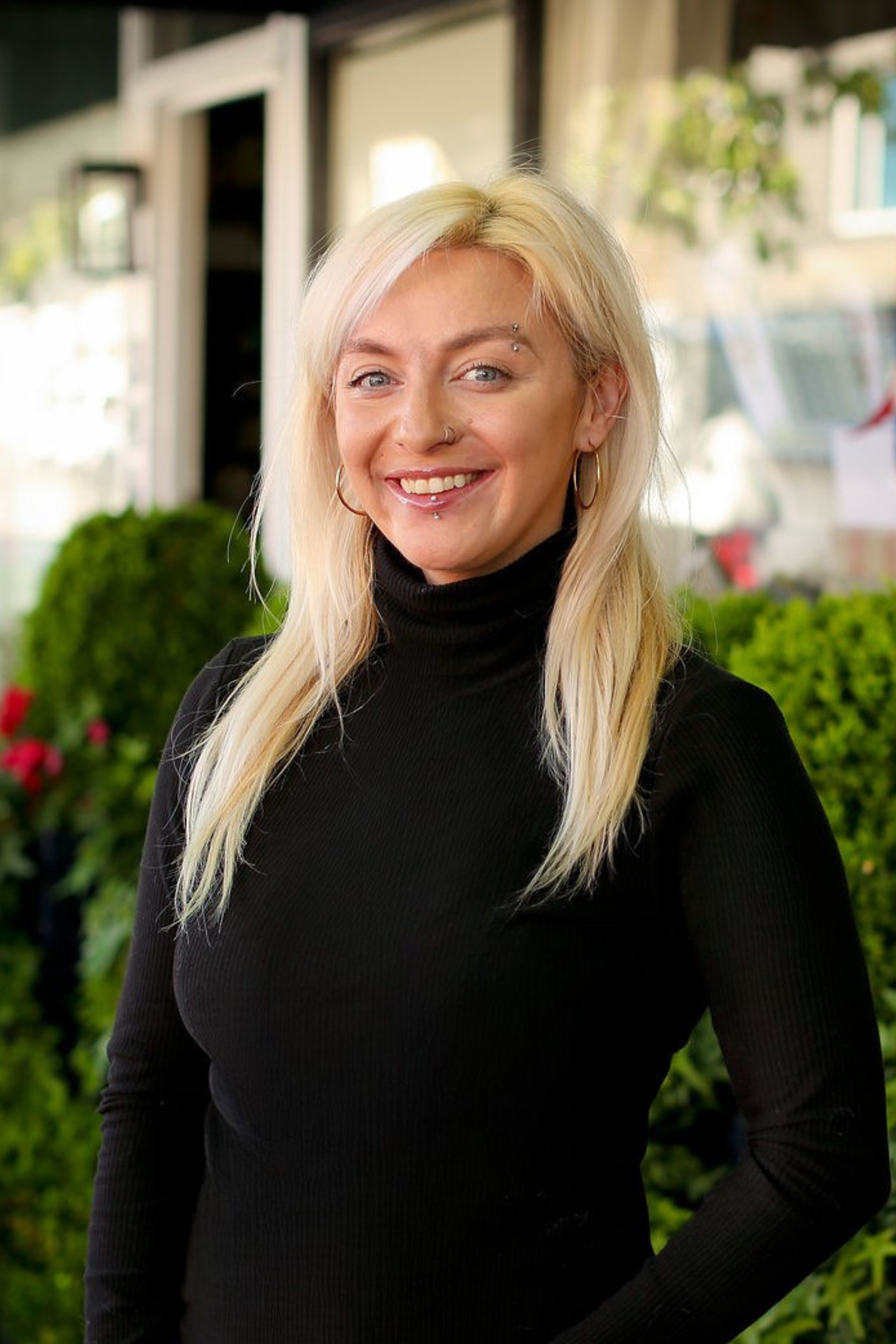 A woman with long platinum blonde hair, wearing a black turtleneck sweater, smiling outdoors in front of greenery and blurred buildings.