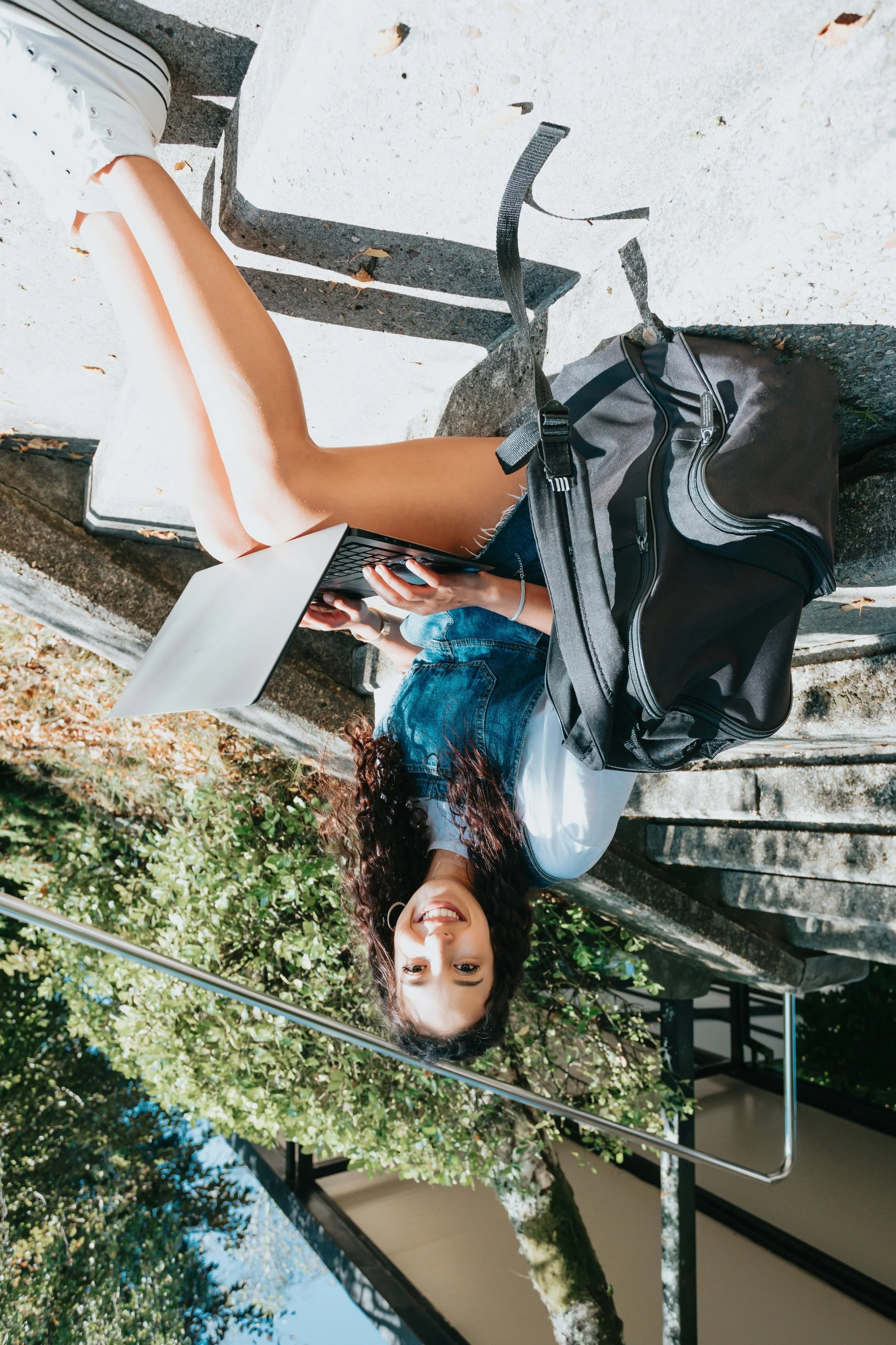 A young woman with curly hair is sitting outdoors on a concrete bench, smiling and looking at the camera. She has a laptop on her lap and a backpack beside her. The background shows green trees and a building, and she appears to be enjoying a sunny day.