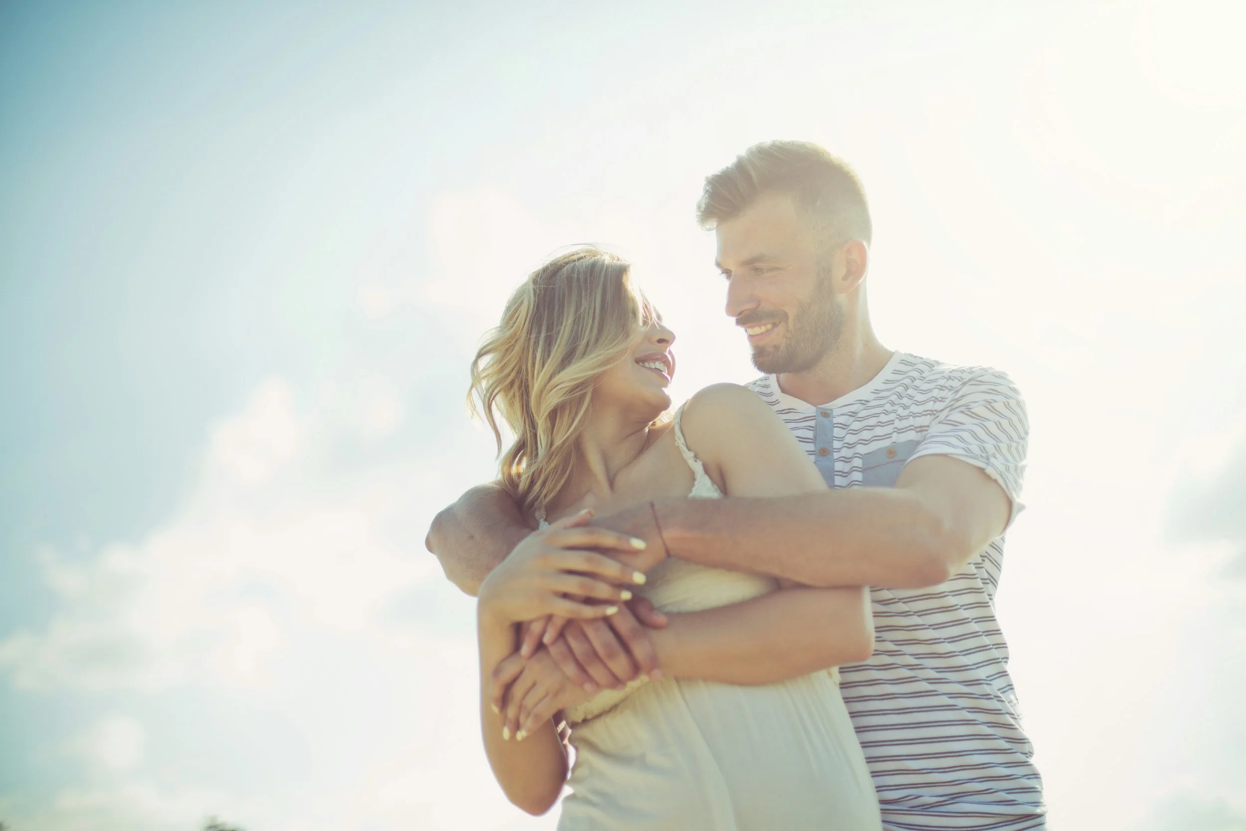 A smiling man and woman are embracing outdoors against a bright sky, with the woman holding a small object in her hand.
