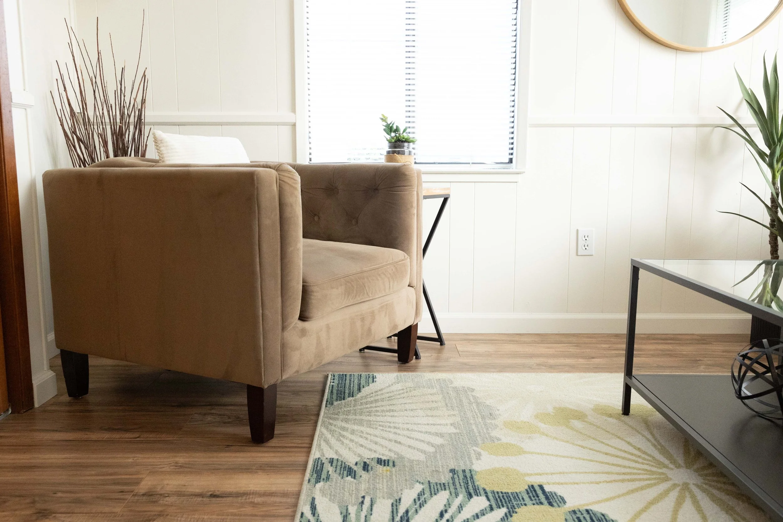 Living room corner with beige upholstered armchair, white pillow, a small side table with a potted succulent plant, a large window with blinds, a wall mirror, plants, power outlet on white paneled wall, wooden floor, and a patterned rug.