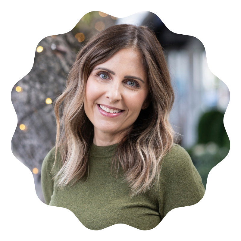 A smiling woman with shoulder-length wavy brown hair, wearing a green sweater, outdoors near a tree with string lights.