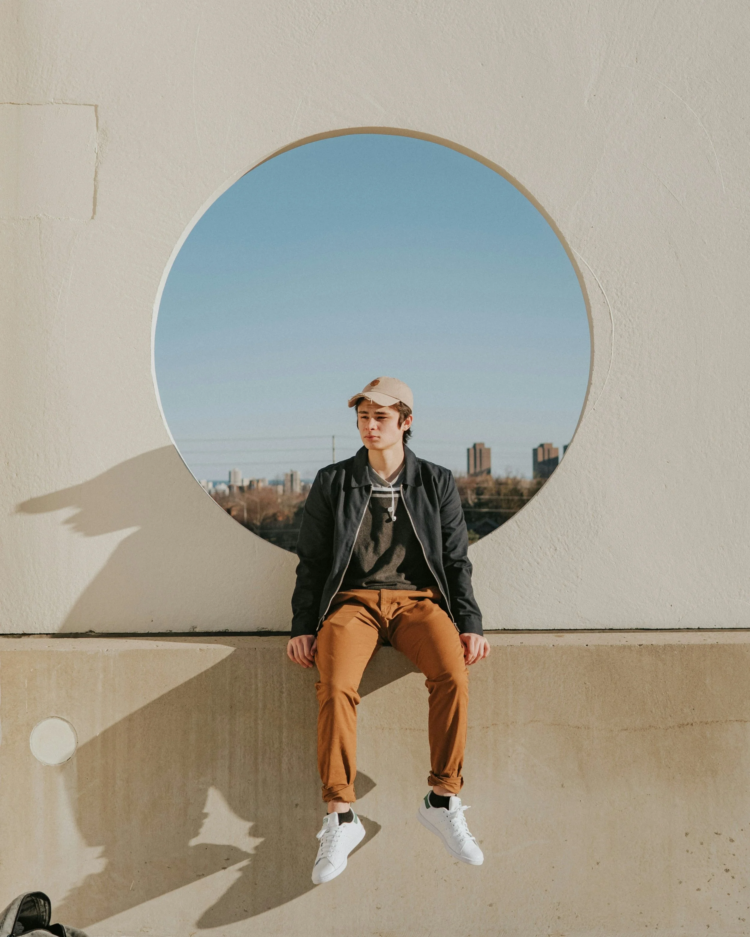 A young man with a tan baseball cap, dark hair, and casual black jacket, sitting on a concrete ledge in front of a geometric wall with a circular opening, overlooking a cityscape with clear skies.