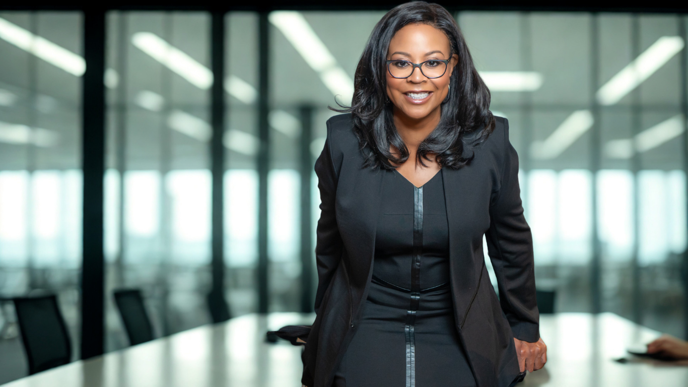 A confident woman in business attire leaning forward in a modern conference room with large windows and chairs.