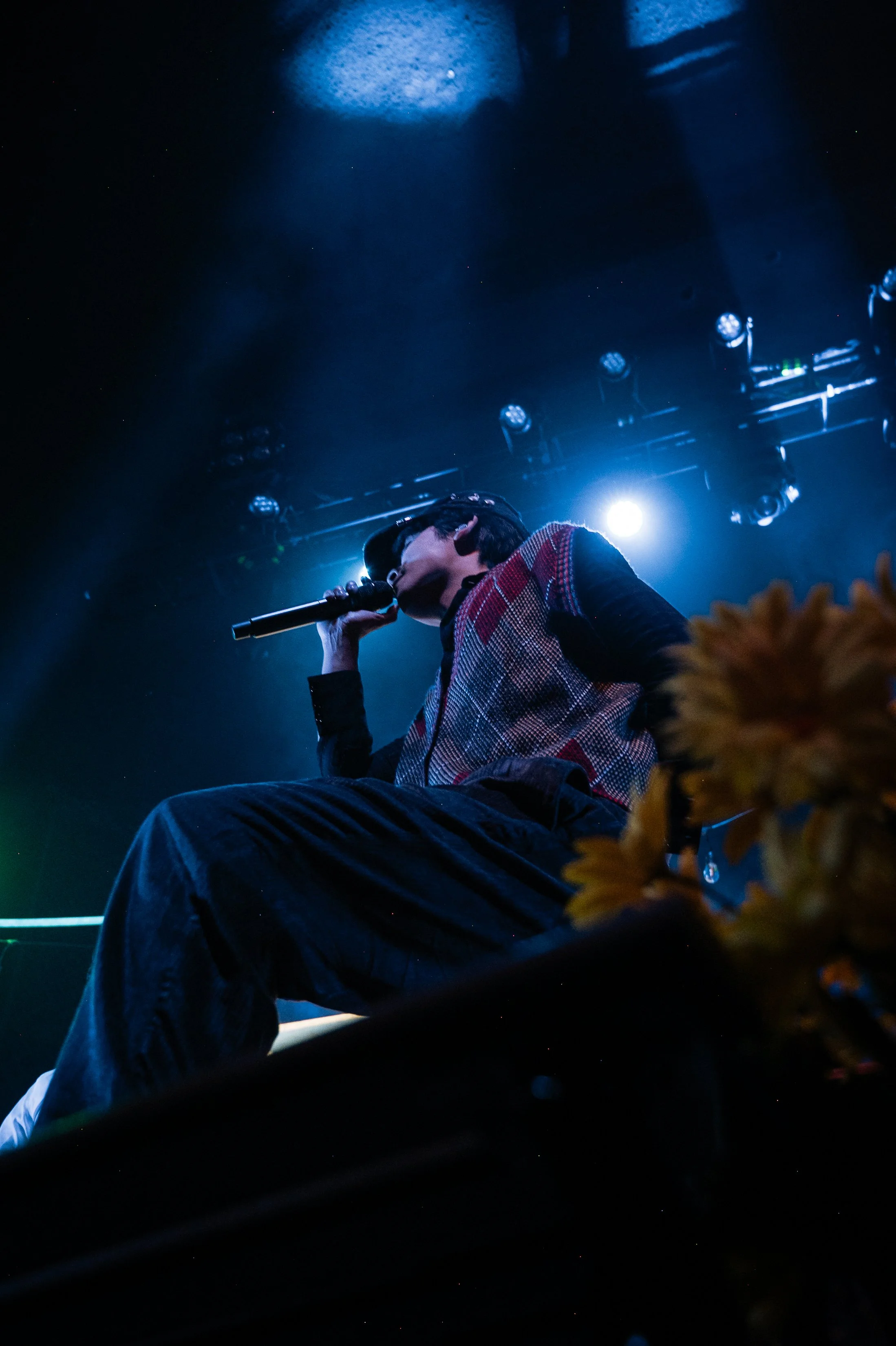 A person sitting on the floor in a concert or stage setting, holding a microphone near their mouth, with bright stage lights shining behind them, and a blurred yellow flower in the foreground.