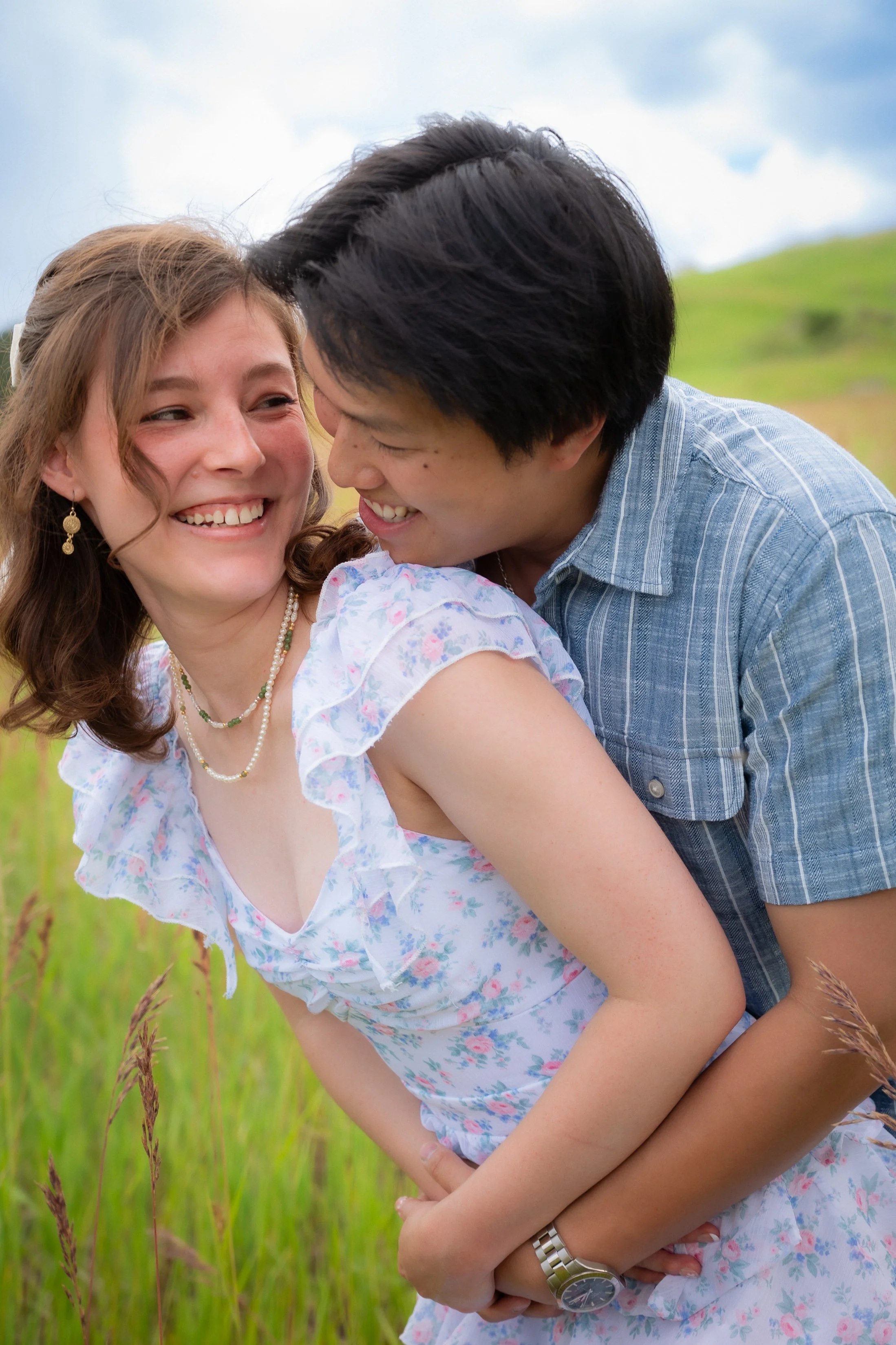 A smiling woman in a floral dress and pearl jewelry being embraced from behind by a smiling man in a blue shirt, outdoors in a grassy field with a cloudy sky.