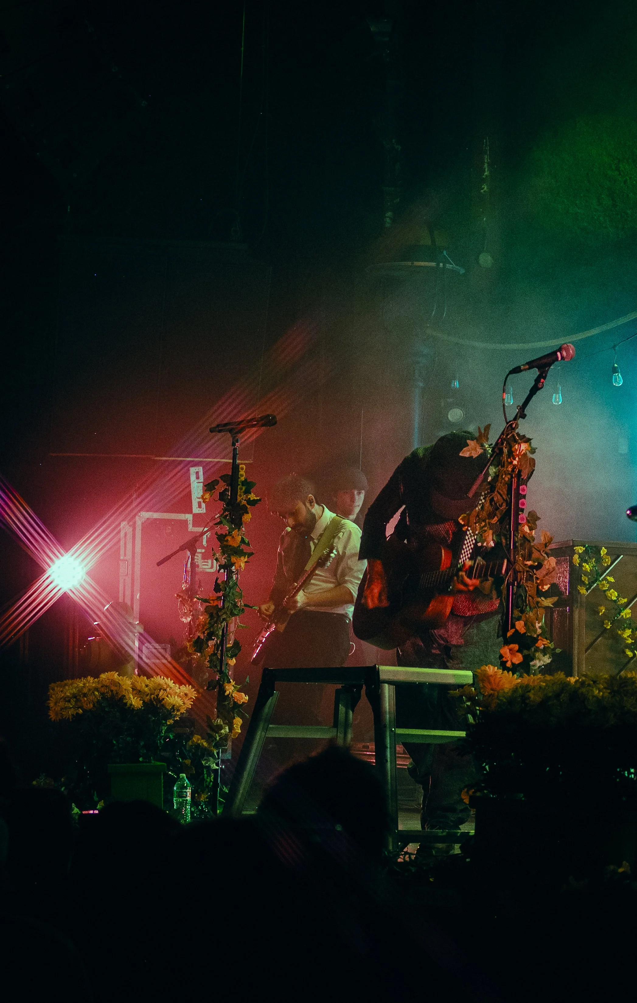 Musicians performing on stage with microphones and guitar, decorated with flowers, under colorful stage lights.