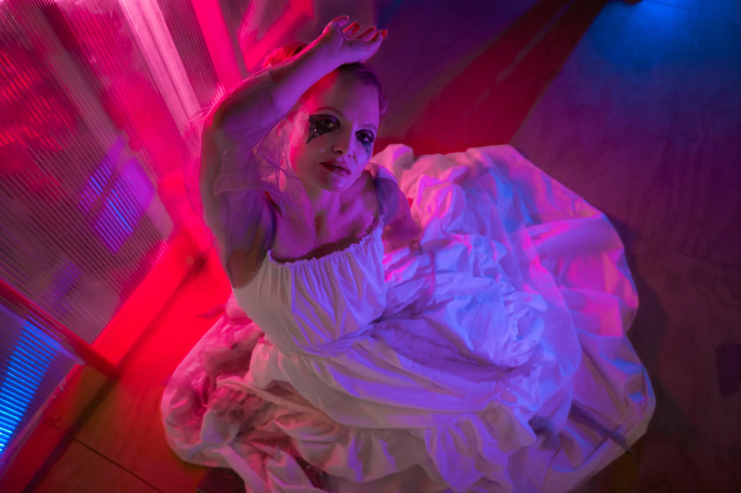 A woman lying on a bed with colorful pink and blue neon lighting, wearing a white dress and makeup that includes dark eyeshadow and lipstick, with a relaxed facial expression.