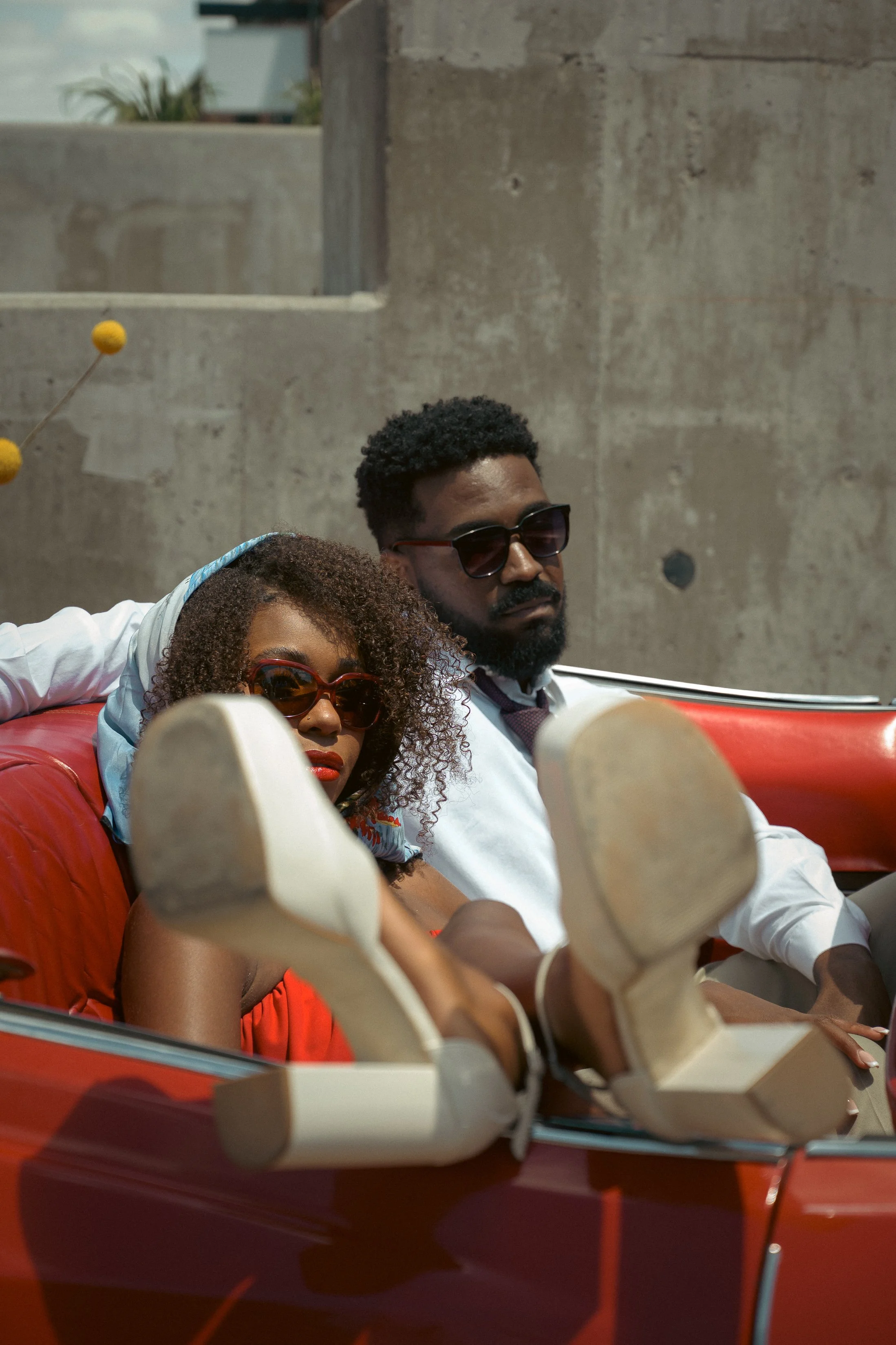 A man and woman sitting together on a red booth seat with concrete walls in the background. They are both wearing sunglasses and are relaxed, with their feet resting on the table.