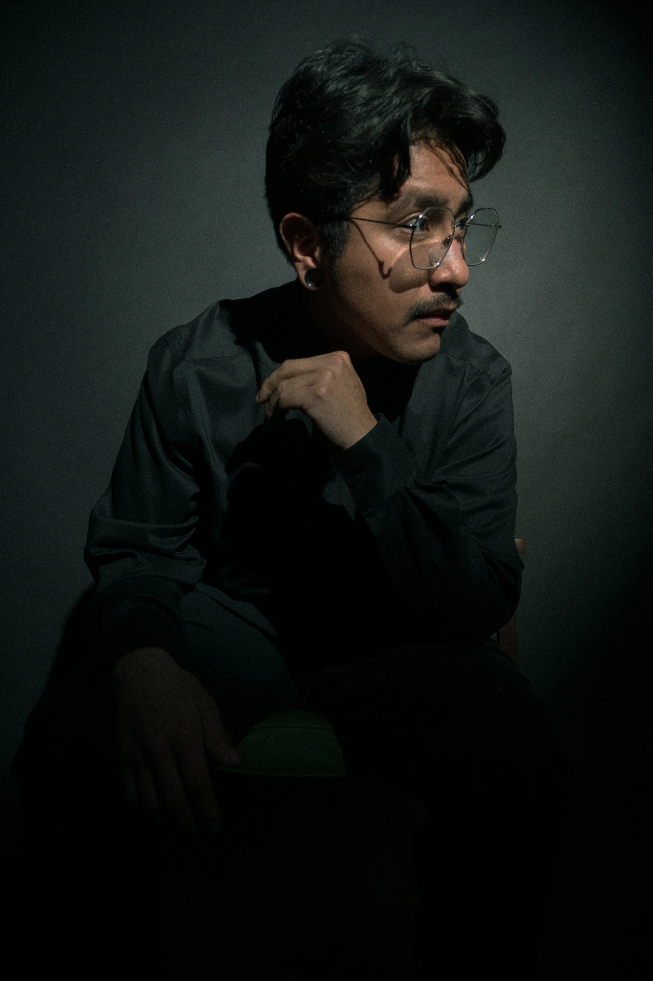 A man with dark hair, glasses, and a goatee, wearing a dark shirt, sitting in low lighting with a contemplative expression.