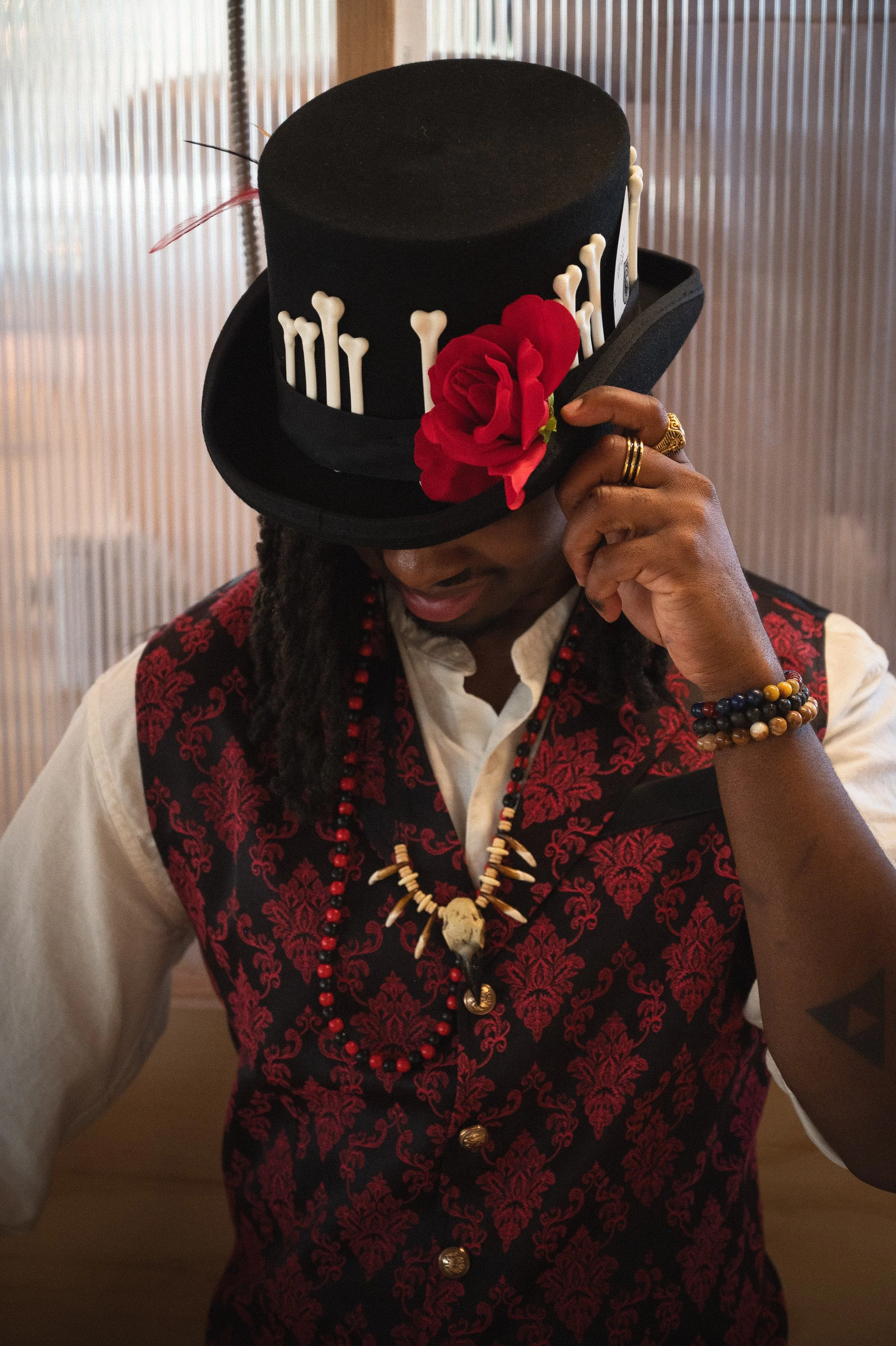 A person wearing a black top hat with a red flower, white bones, and a black ribbon, holding the brim with one hand and wearing rings and beaded necklaces, dressed in a white shirt and a red and black embroidered vest.
