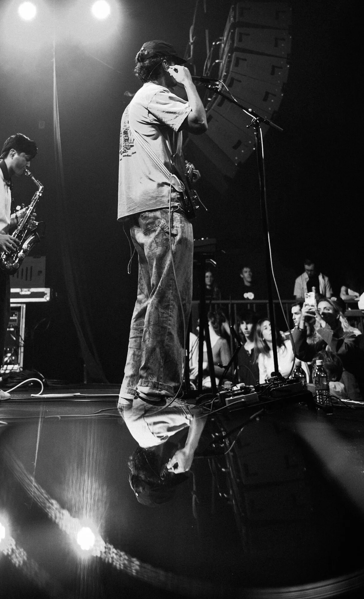 Black and white photo of a musician on stage, wearing a hat, holding a microphone, and singing or speaking. Another musician is visible playing a saxophone, and there is a crowd in front of the stage with some people taking photos.