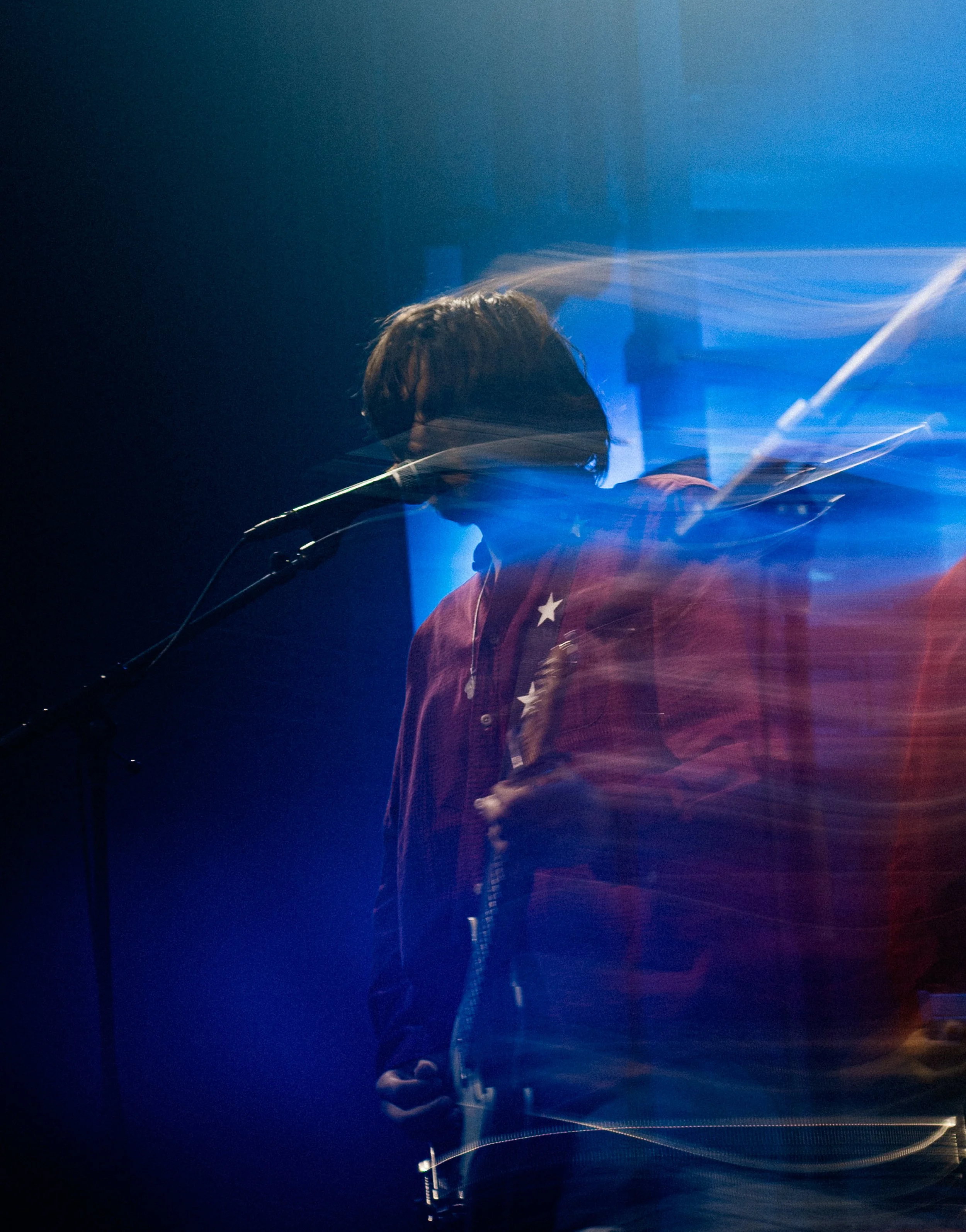 A person with curly hair and a red jacket playing an electric guitar on stage, illuminated by blue stage lights.