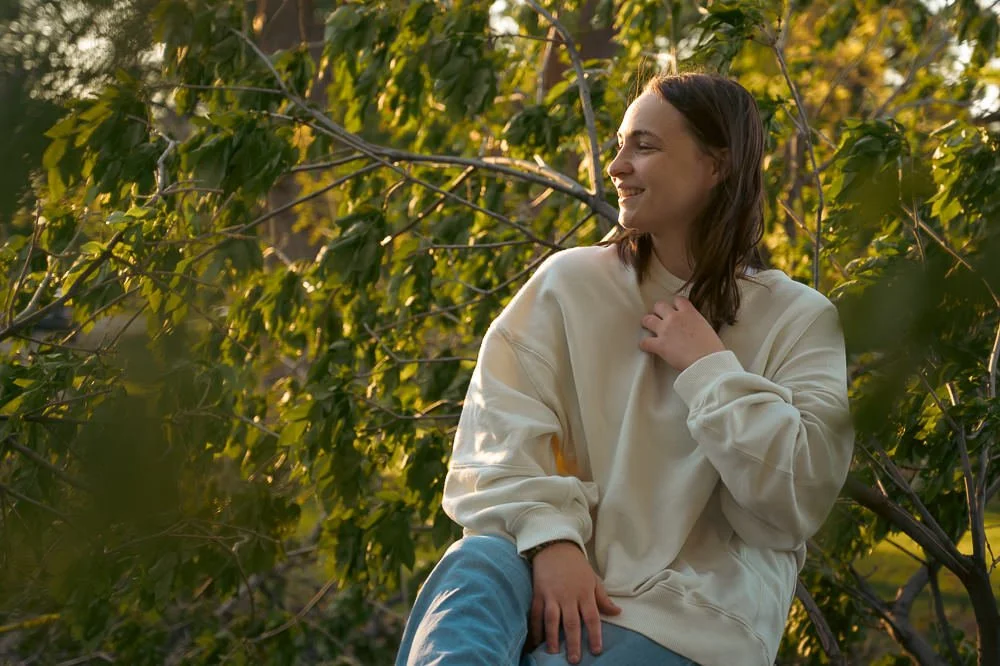 A young woman sitting on a tree branch surrounded by green leaves during sunset, smiling and wearing a cream-colored sweatshirt.