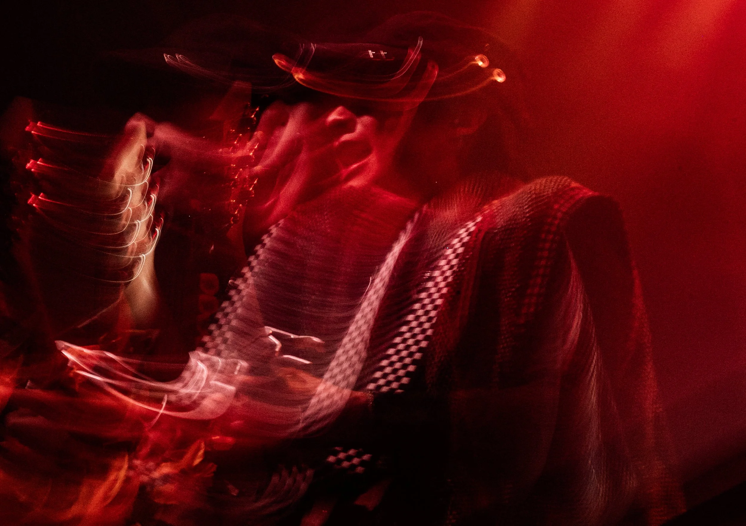 A person wearing a hat and checkered shirt, with their hand near their face, illuminated in red light with abstract streaks and light trails.