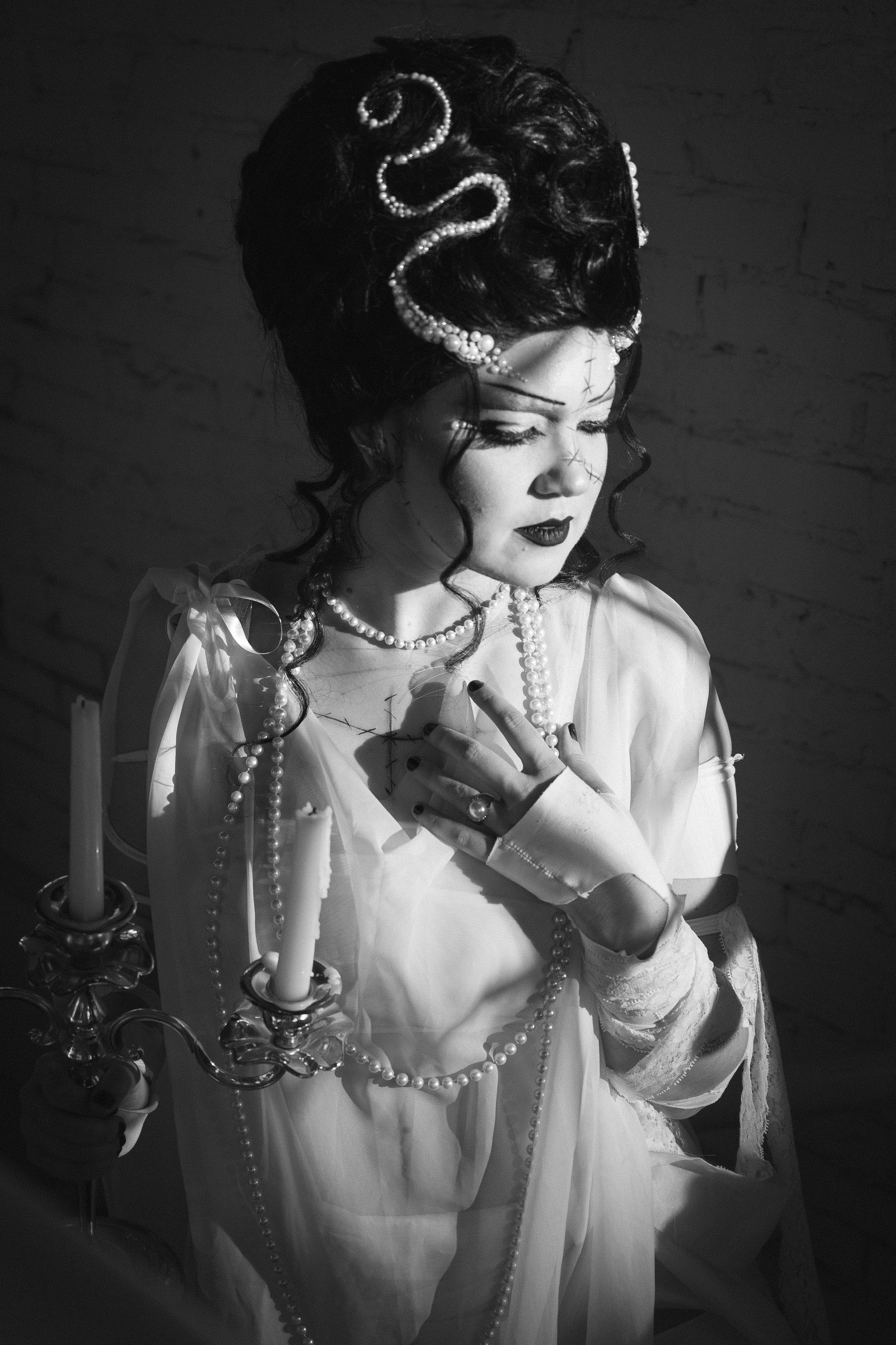 A woman dressed as a bride with dark curls, pearl jewelry, and a white gown, holding a candelabra, with a brick wall in the background.