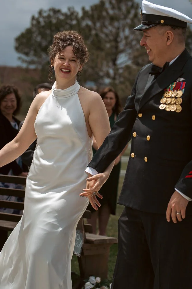 Couple holding hands and smiling, the woman in a white wedding dress and the man in a formal military uniform, outdoors with guests in the background.