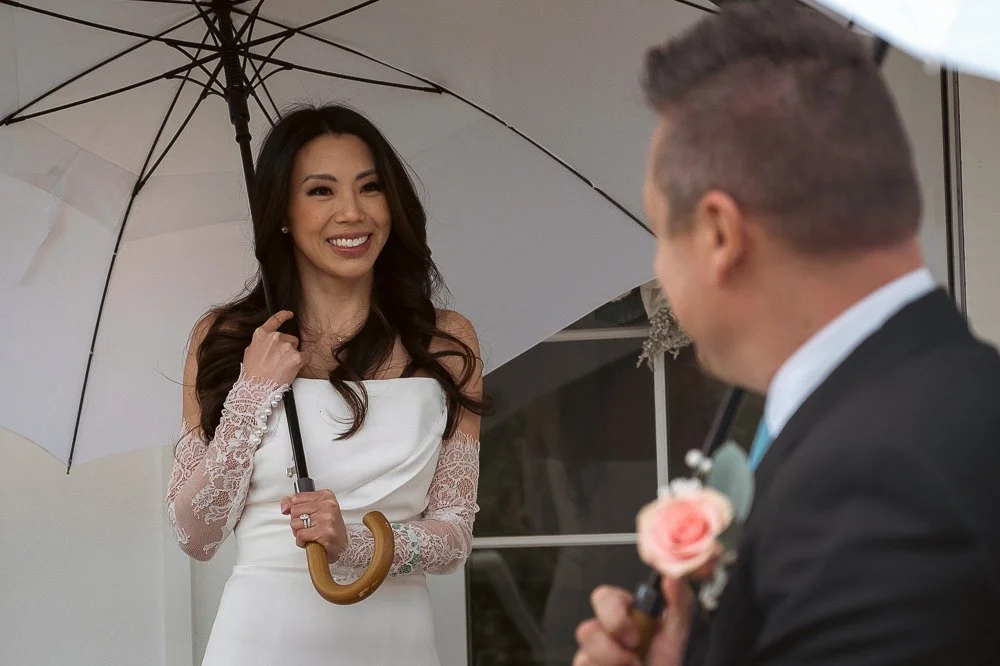 A woman in a white dress with lace sleeves holding an umbrella, smiling at a man in a suit who is also holding an umbrella during a rainy day.