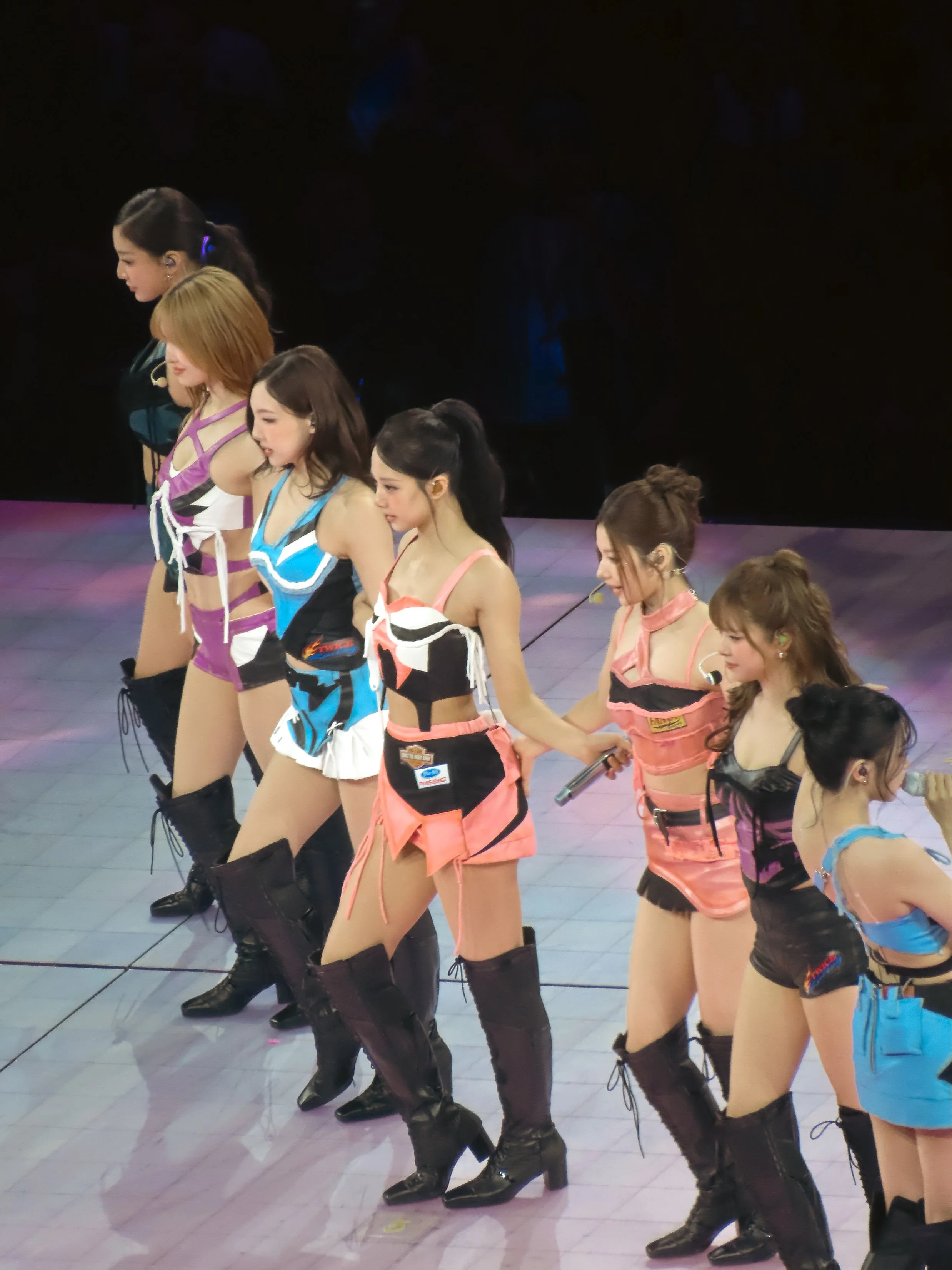 Six young women performing on stage, wearing colorful outfits and black boots, with some holding microphones.