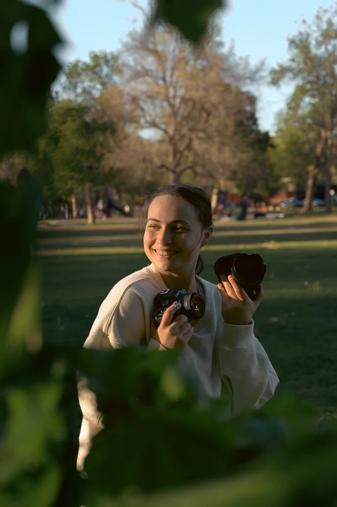 A young woman smiling and holding two cameras outdoors in a park with trees and a grassy area.
