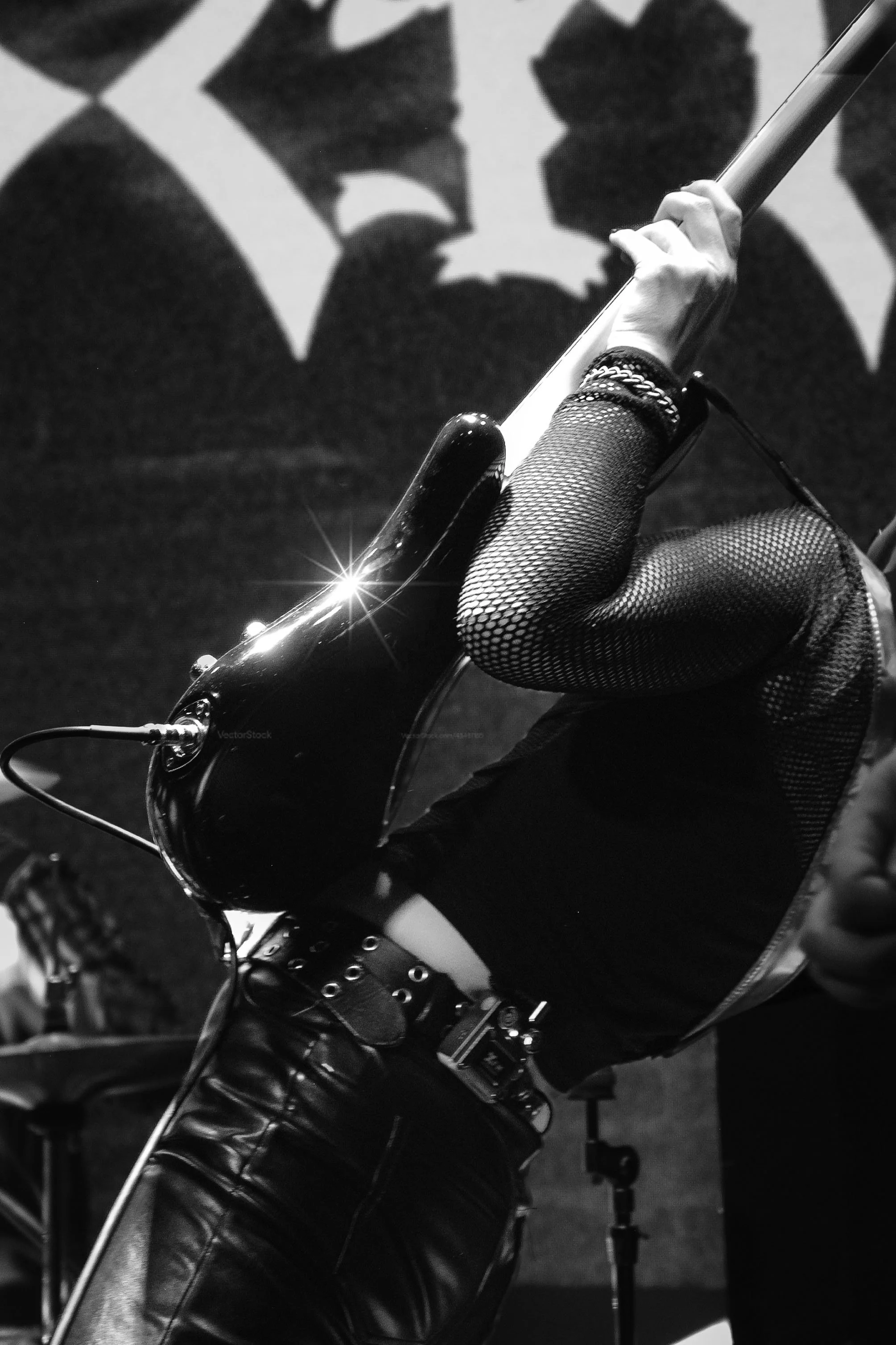 Close-up of a person wearing fishnet stockings and a leather jacket, holding a guitar neck. Behind them, a graphic of a woman's face in contrast, appears on a wall.