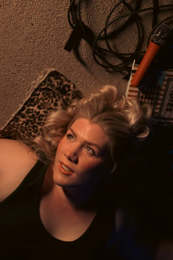 A woman with blonde curly hair lying on a leopard print rug, wearing a black top, in a dimly lit room with miscellaneous cables and objects nearby.