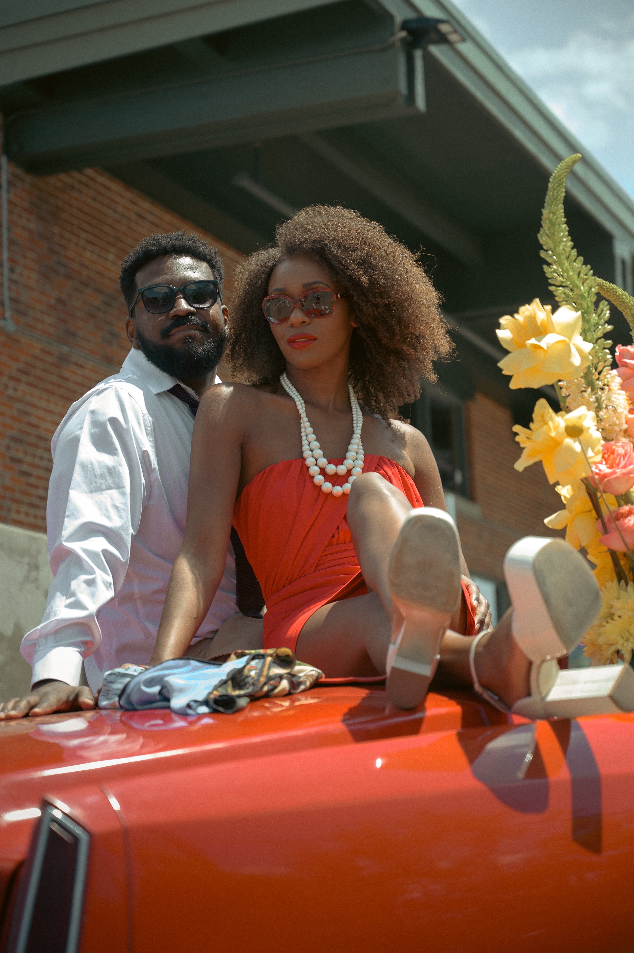 A man and woman sit on a red car decorated with flowers. The woman wears a red dress, pearl necklace, and sunglasses, with her legs stretched out on the hood. The man wears a white shirt and sunglasses, leaning on the car.