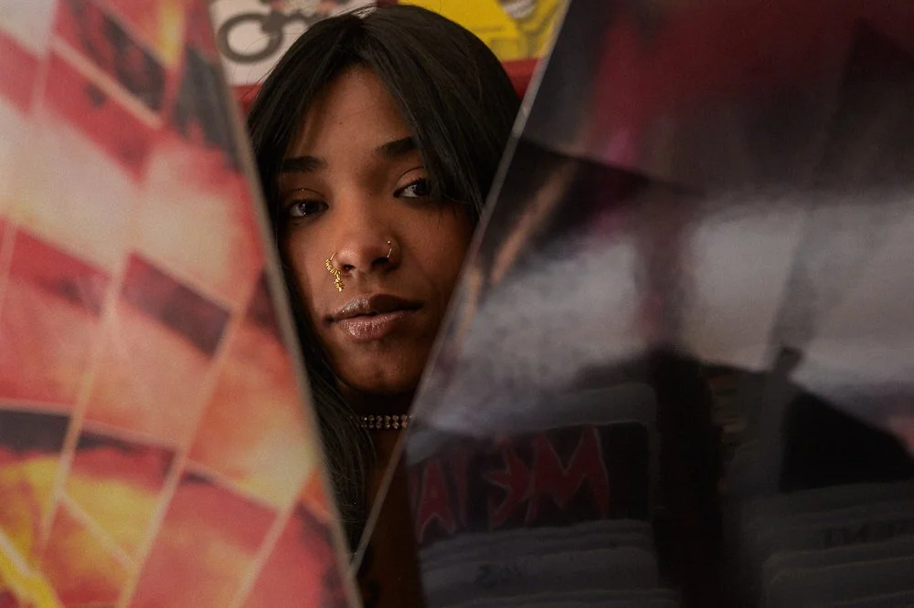 A young woman with medium skin tone and long dark hair looking through a gap between two objects, with these objects partially obstructing her view. She has a nose piercing and wears a choker necklace.