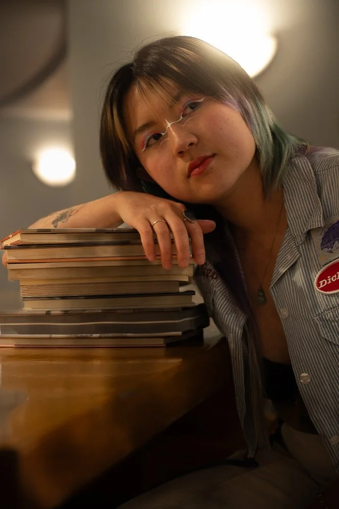 A young woman with short, multi-colored hair, wearing glasses and a striped button-up shirt, resting her head on her arm next to a tall stack of books on a wooden table.