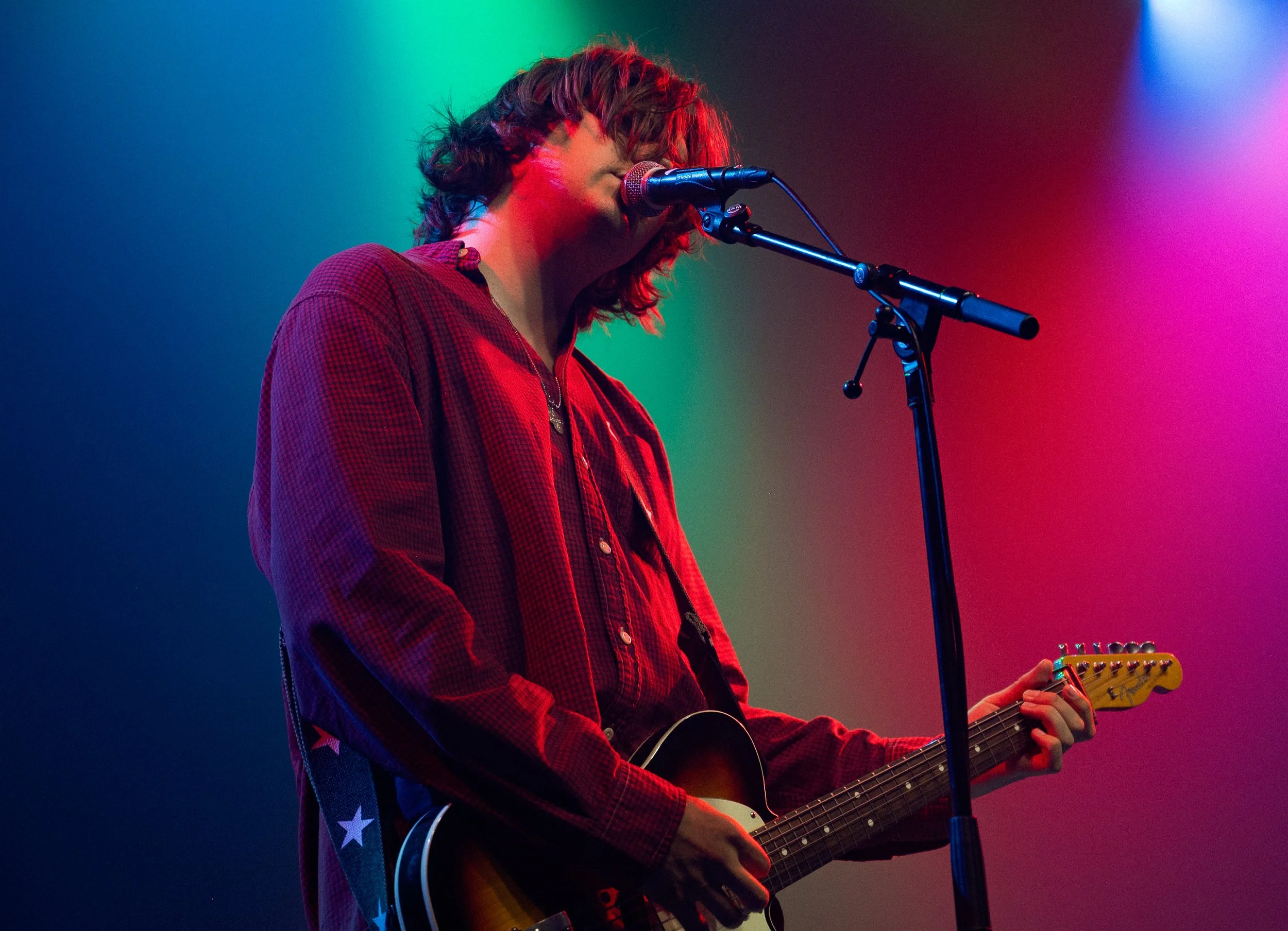 A person with curly hair wearing a red checkered shirt playing an electric guitar and singing into a microphone on stage with colorful stage lighting.