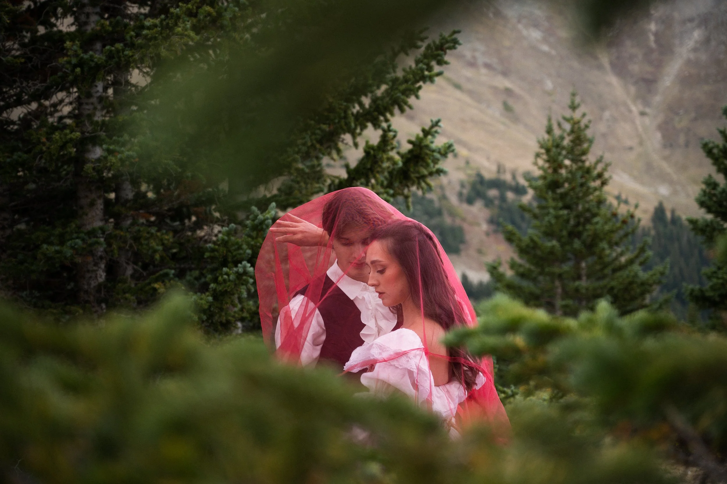 A couple dressed in vintage style, the man with a dark vest and white shirt, and the woman with long hair and a pink veil, standing outdoors among trees with mountains in the background.