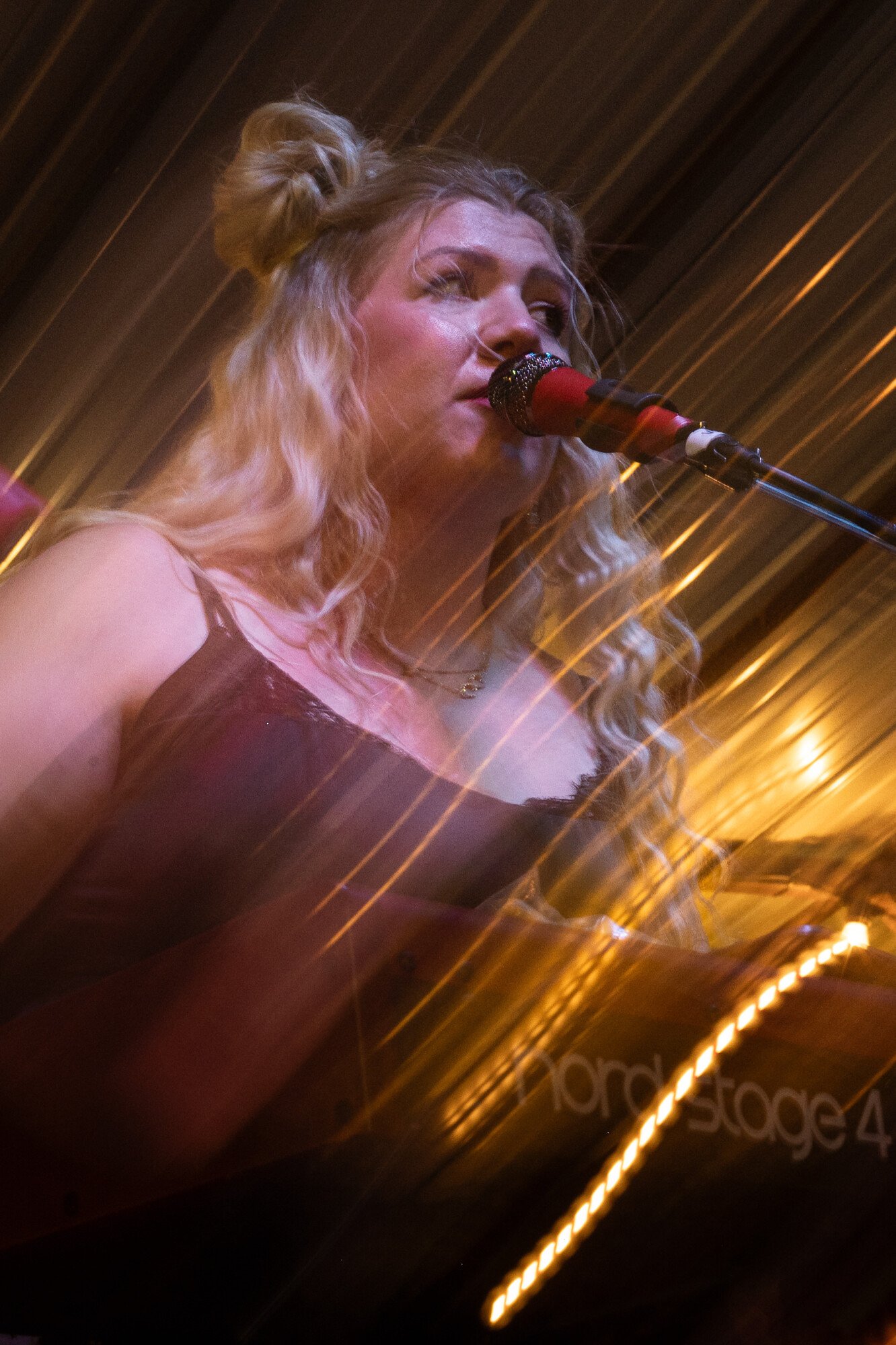 A woman with long, wavy blonde hair wearing a sleeveless dress, singing into a microphone on a stage with warm lighting and wooden paneling in the background.