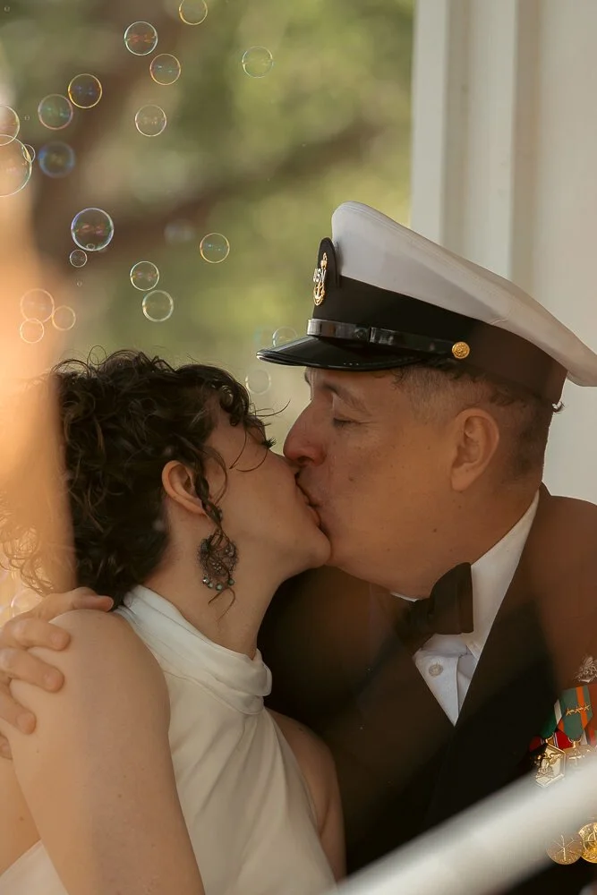 A couple sharing a kiss: woman with curly hair and earrings, man in military uniform with a white hat and medals, in front of a window with sunlight.