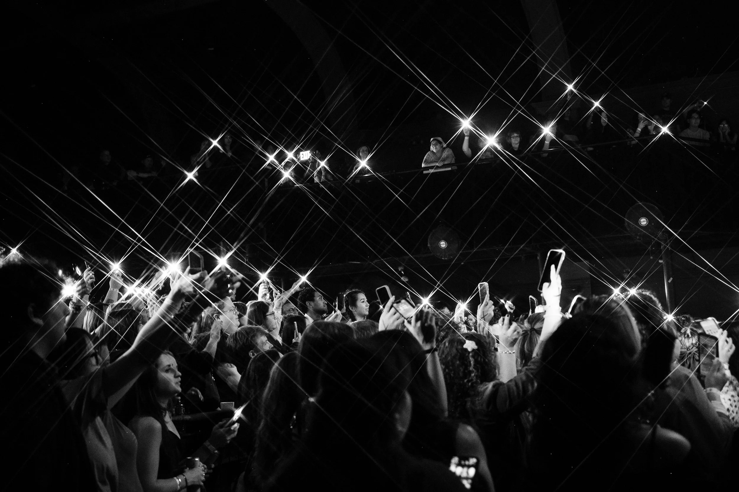 Crowd of people attending an event, many taking photos with their phones, with bright lights creating starburst effects in the background.