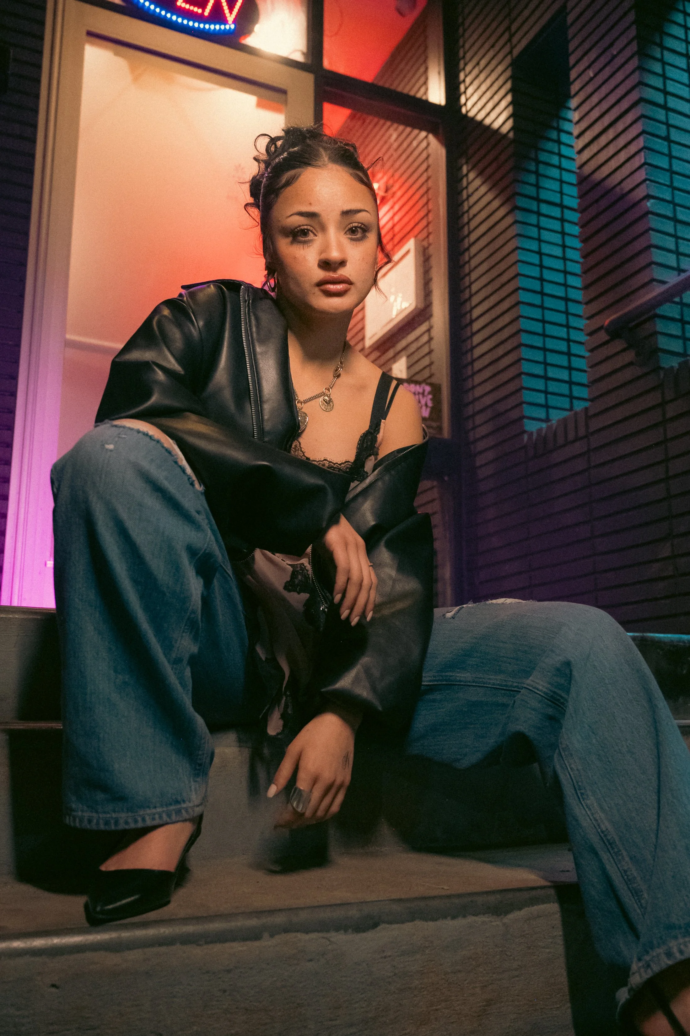 A young woman sitting outdoors at night, wearing a black leather jacket and ripped jeans, with a neon sign that says 'LIVE' in the background, and colorful lighting reflecting on the scene.