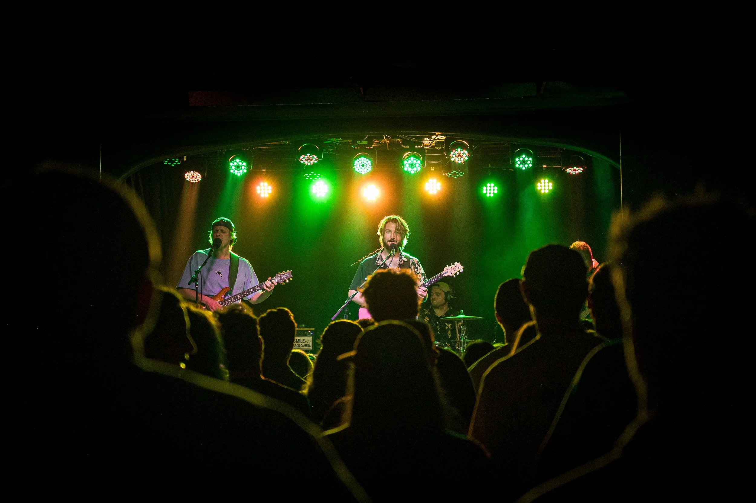 A band performing on stage with colorful green, yellow, orange, and red stage lights, and the audience in the foreground.
