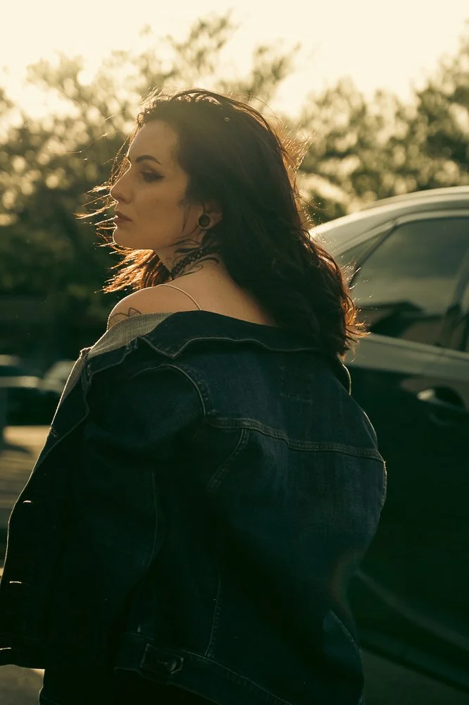 A woman with dark brown hair standing outdoors near a parked car during sunset, wearing a dark denim jacket.