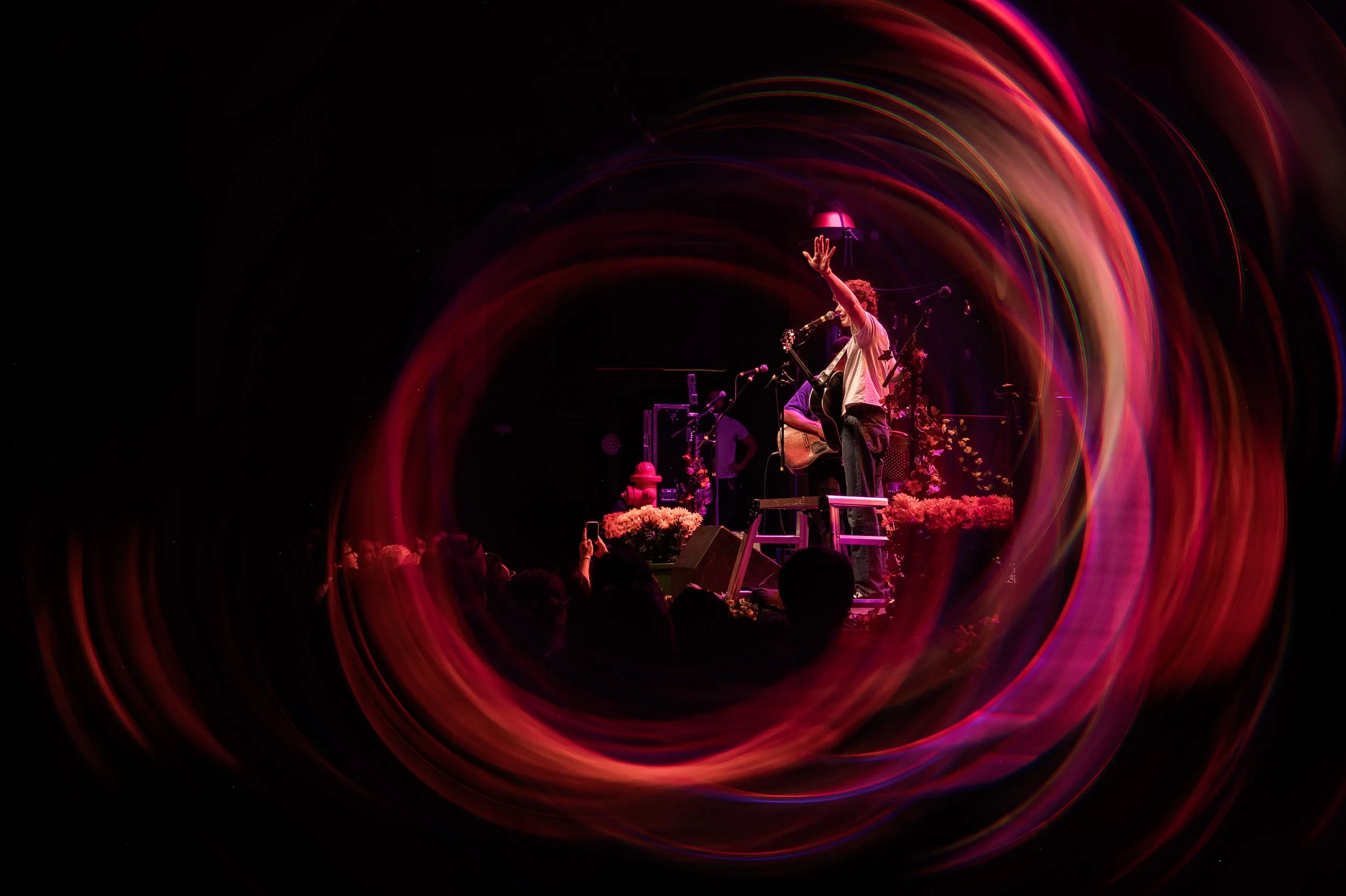 Performer on stage playing guitar, reaching out toward audience, with colorful light trails swirling around in a dark setting.