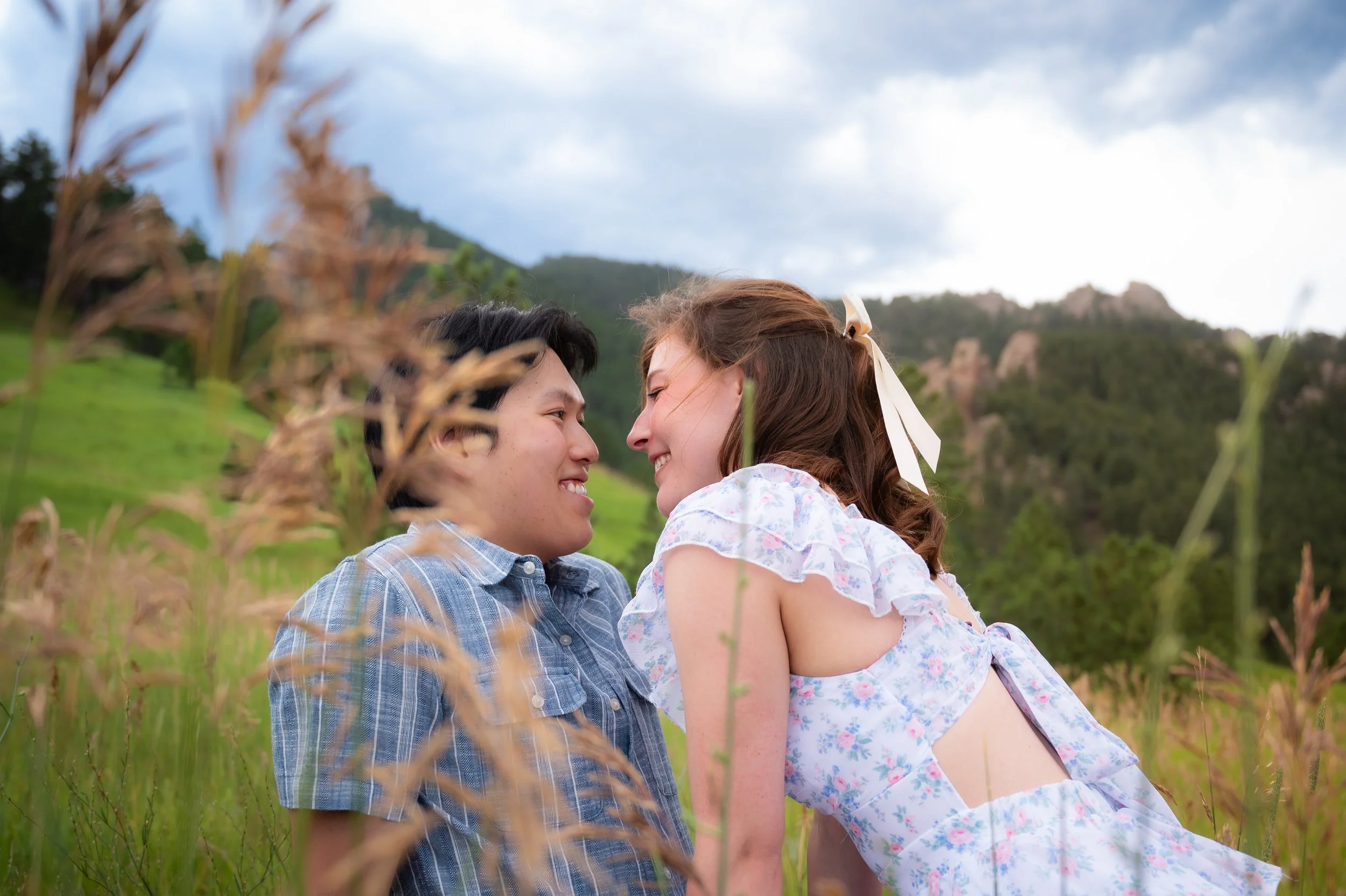 A young couple, sitting close together outdoors in a field of tall grass, smiling and facing each other with their foreheads touching. The background features green hills and a cloudy sky.